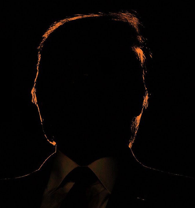 Backlit view of a man's head and shoulders. He is wearing a suit and tie. His facial features are in shadow and not visible. The light behind him casts his hair around his head in an orange glow.