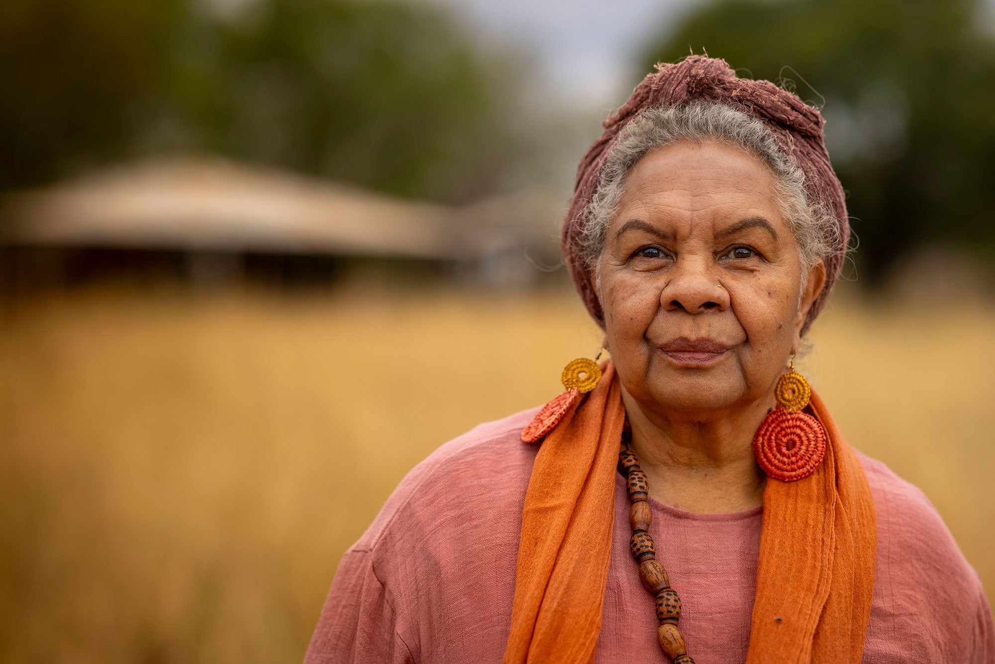 Anne stands facing with the viewer with a serious expression. A dwelling is out of focus in the background. The background is a blur of rich yellow and green. Anne is wearing large circular colorful earrings. Her hair is tidily tied up in a pink turban.
