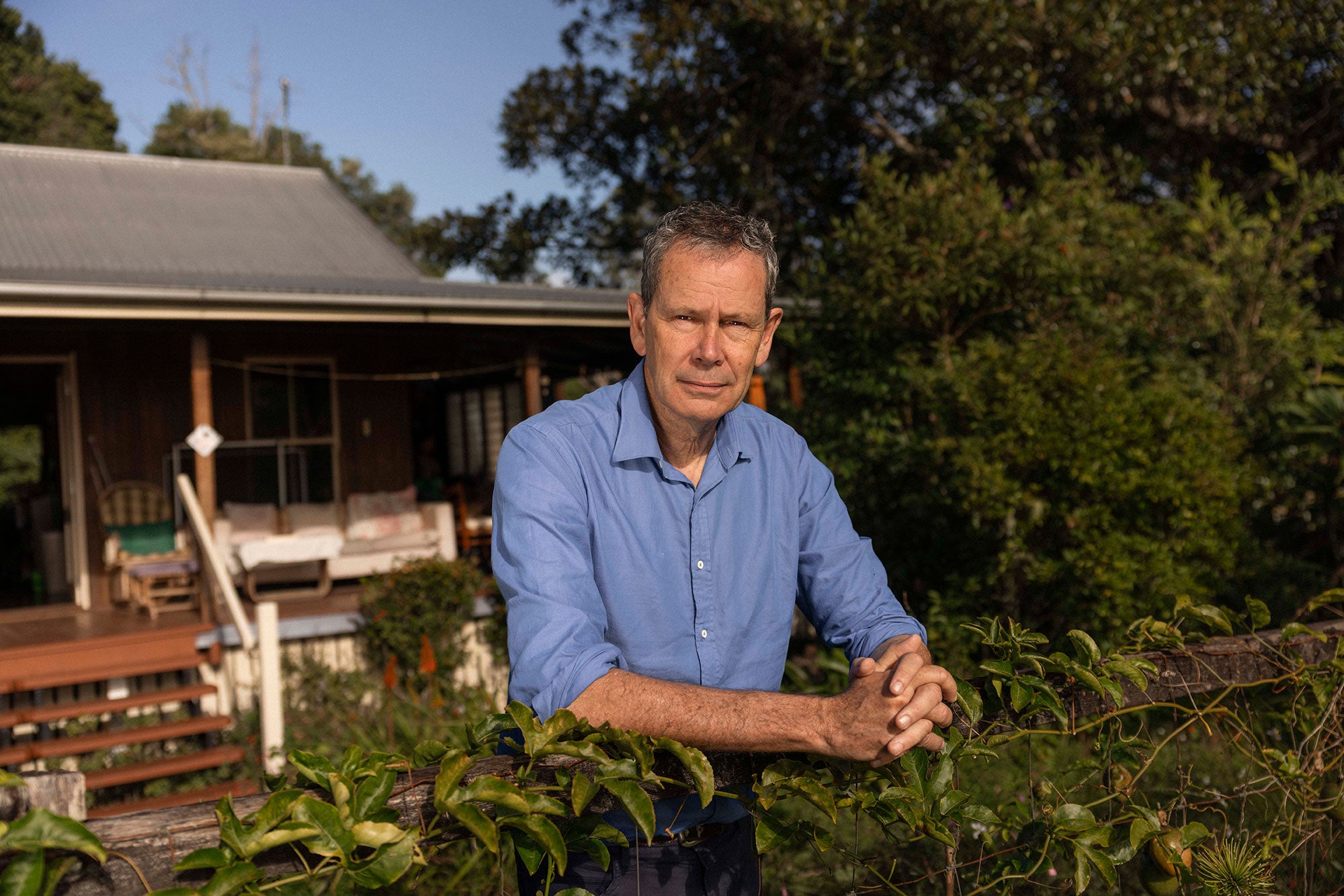 Barry stands in his front yard, resting his forearms on a fence with his hands clasped in front of him. A passionfruit vine grows entwined along the fence. Barry looks directly at the viewer. It is a bright sunny day, with the sun shining on Barry and blue sky visible in the distance. Barry's home is visible behind him, with a large tree to the side. There are comfortable chairs lining the front porch, with six steps leading up to the house from the ground level.