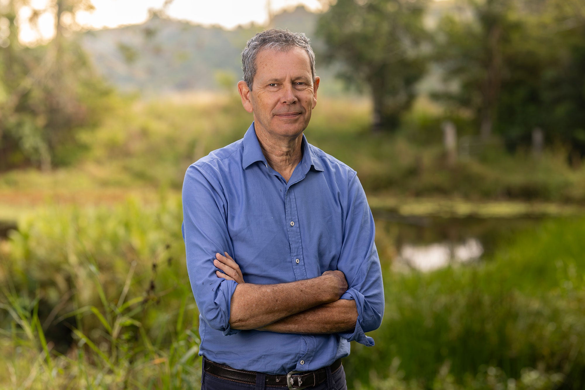 Barry, with a slight, serious smile, stands directly facing the viewer with his arms crossed in front of him. He is standing in a green, open field with tall grasses and the background out of focus. Barry is wearing a collared, buttoned-up long sleeve shirt, with the sleeves rolled up to his elbows.