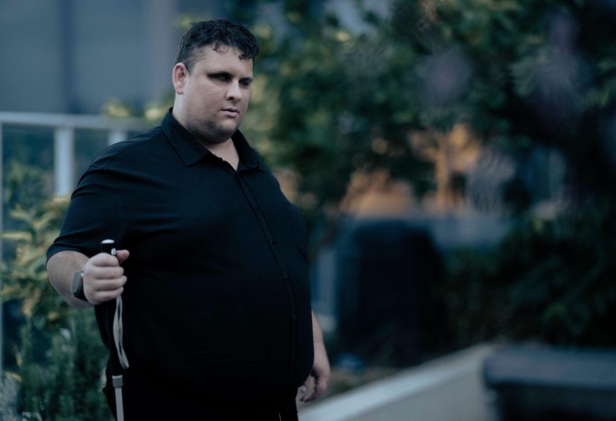 Brendon, standing with plants and rooftop architecture in the background (Blurred). A medium shot. You see him from waist upwards. Brendon is slightly to the left and is turned to his left looking thoughtfully to the side. He is holding his walking cane to the right side. It is late in the afternoon. The photo has a golden light.