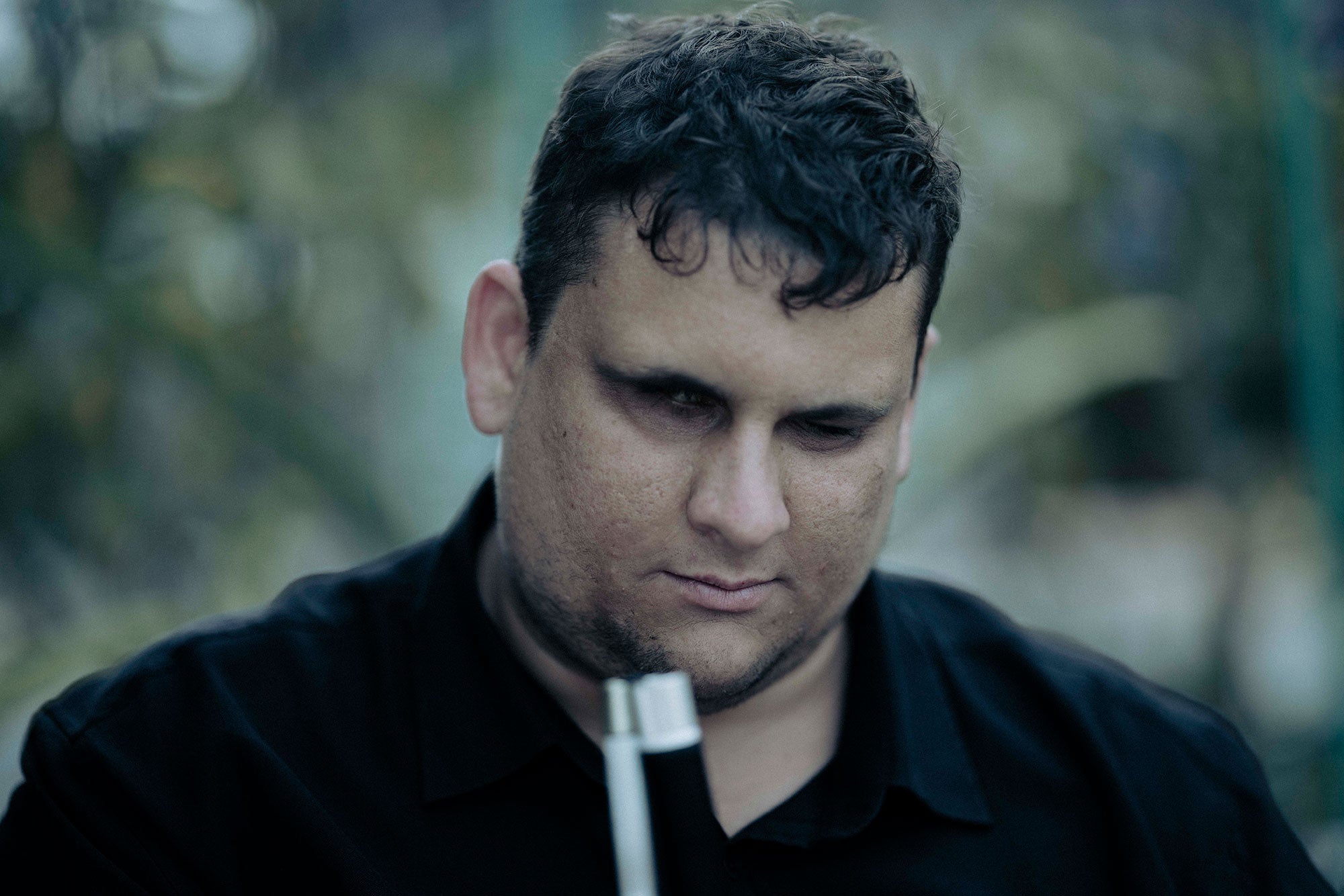 Brendon, seated with plants in the background (Blurred). A close-up. the photo is focussed on his face. Brendon is looking down at the ground with a thoughtful expression. You can see the top of his walking cane which he is holding. It is late in the afternoon. The photo has a golden light.