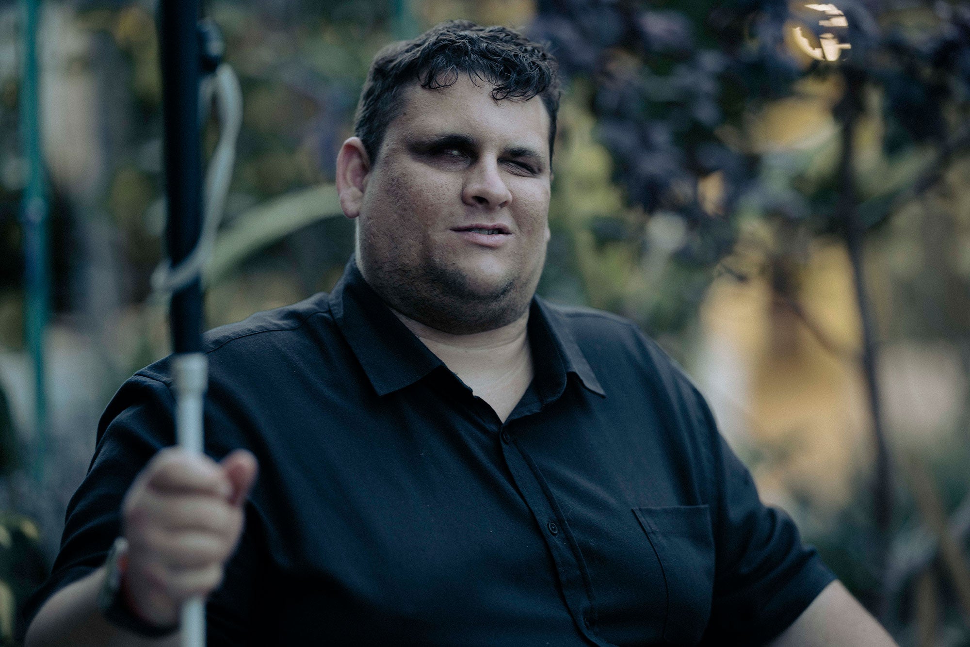 Brendon, seated, with plants in the background (Blurred). It is a medium close-up. You see him from mid-chest upwards. Brendon is looking directly at the camera, with a slight smile and he is holding his walking cane to the side. It is late in the afternoon. The photo has a golden light which makes it look cinematic.