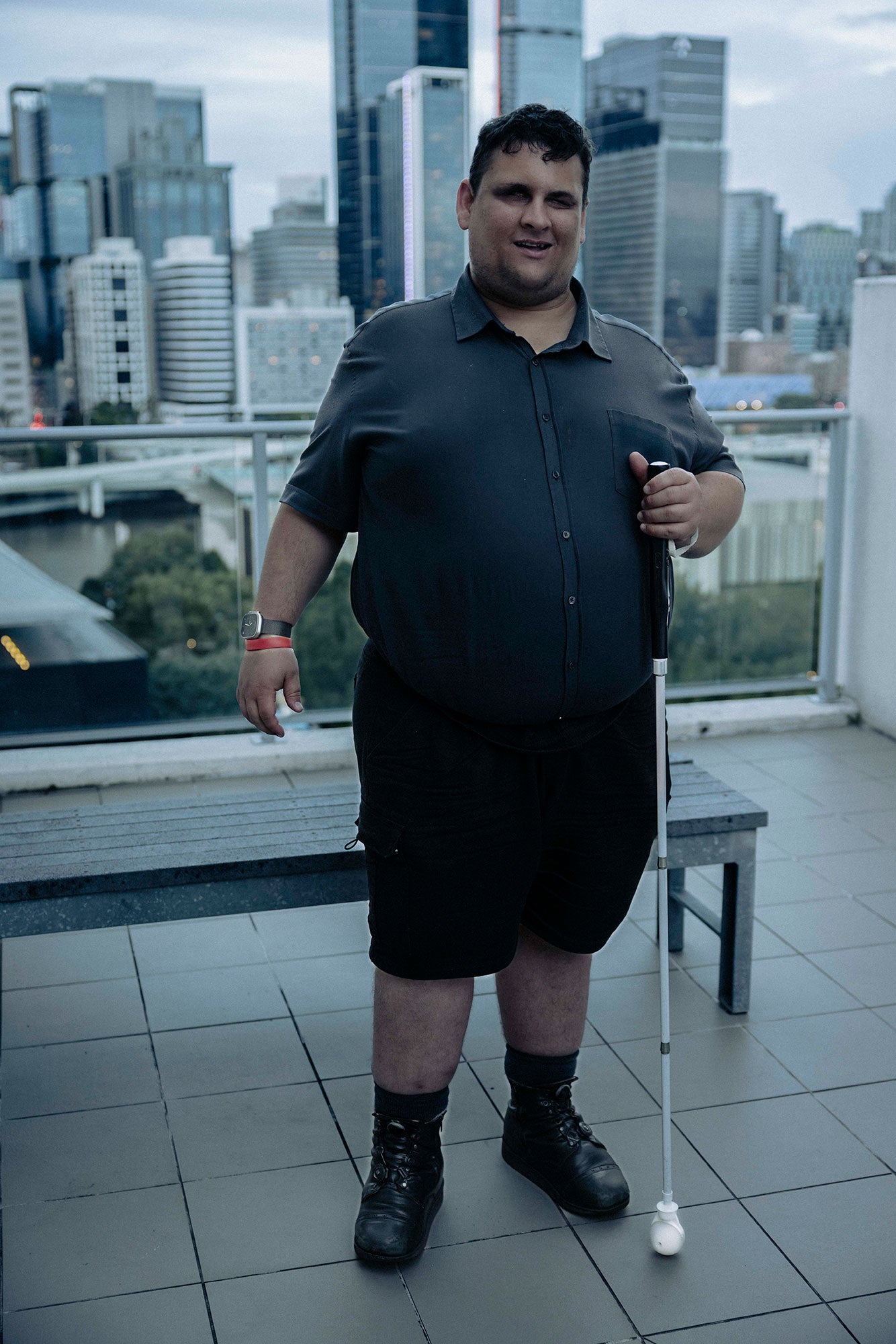 Brendon, standing, with the Brisbane cityscape in the background. It is a full-body shot. You see him fully standing. Brendon is looking directly at the camera and has a slight smile. He is holding his walking cane to the side. It is late in the afternoon. The photo has a blue light which makes the photo look cinematic.