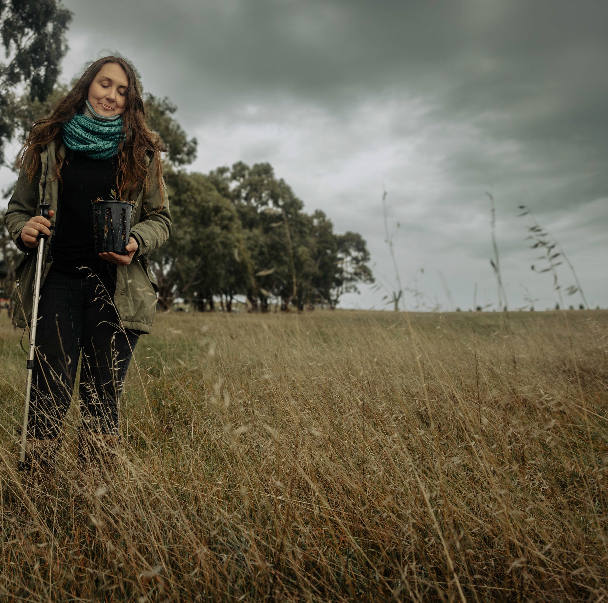 Cat stands in an open field of tall, yellowish-brown grass. She is looking down, with a melancholic, slight smile and her eyes partly closed, at a plant in a black pot that she's holding up to waist level with her left hand. Her right hand is holding a hiking pole. The sky is overcast with dark grey clouds. A line of large trees with thick foliage are in the distant background.