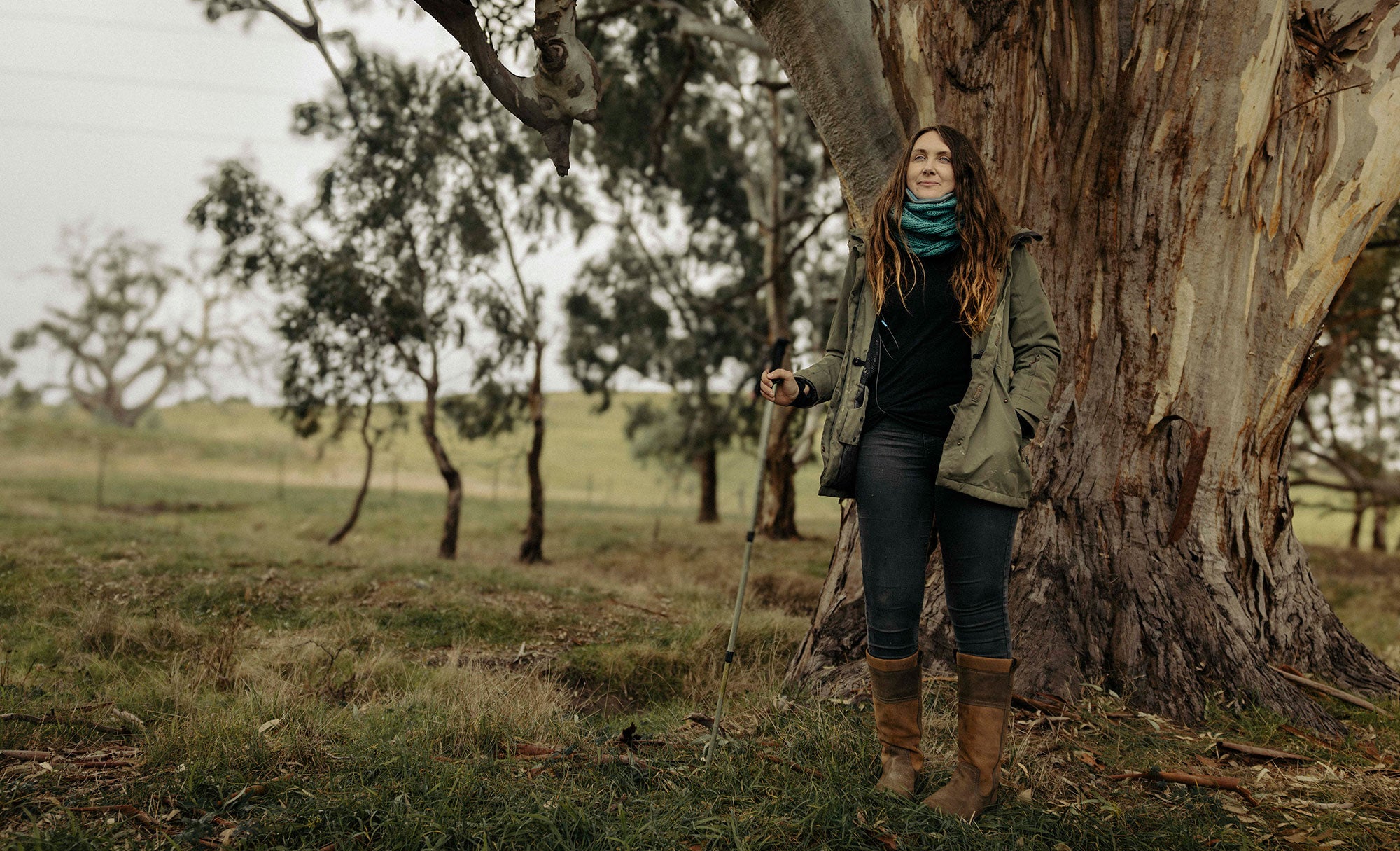 Cat stands in a grassy field in front of a tree with an impressively thick trunk. Light brown pieces of bark are peeling off the trunk, revealing lighter brown bark. several other smaller trees with thin trunks are in the background. Cat is holding a hiking pole, and looks slightly off to the side, with a slight smile.