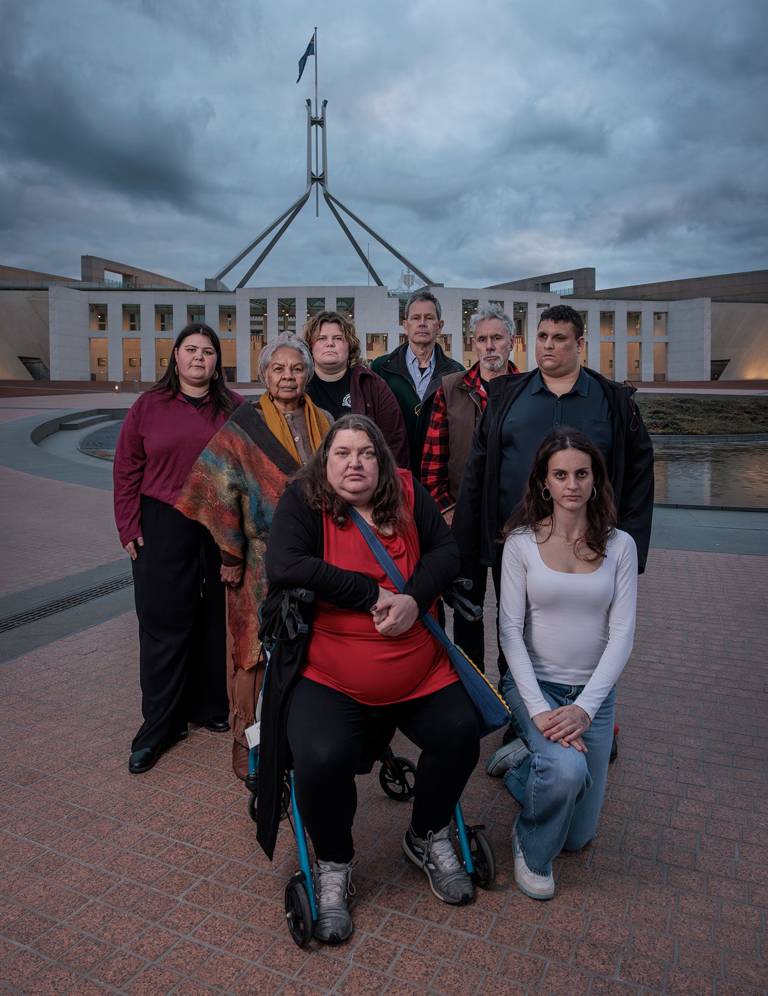 A group of 10 people, looking directly at the viewer, stand in front of Parliament House in Canberra. There are dark clouds overhead, and the soft golden lights shine up onto the front of the building from its base.