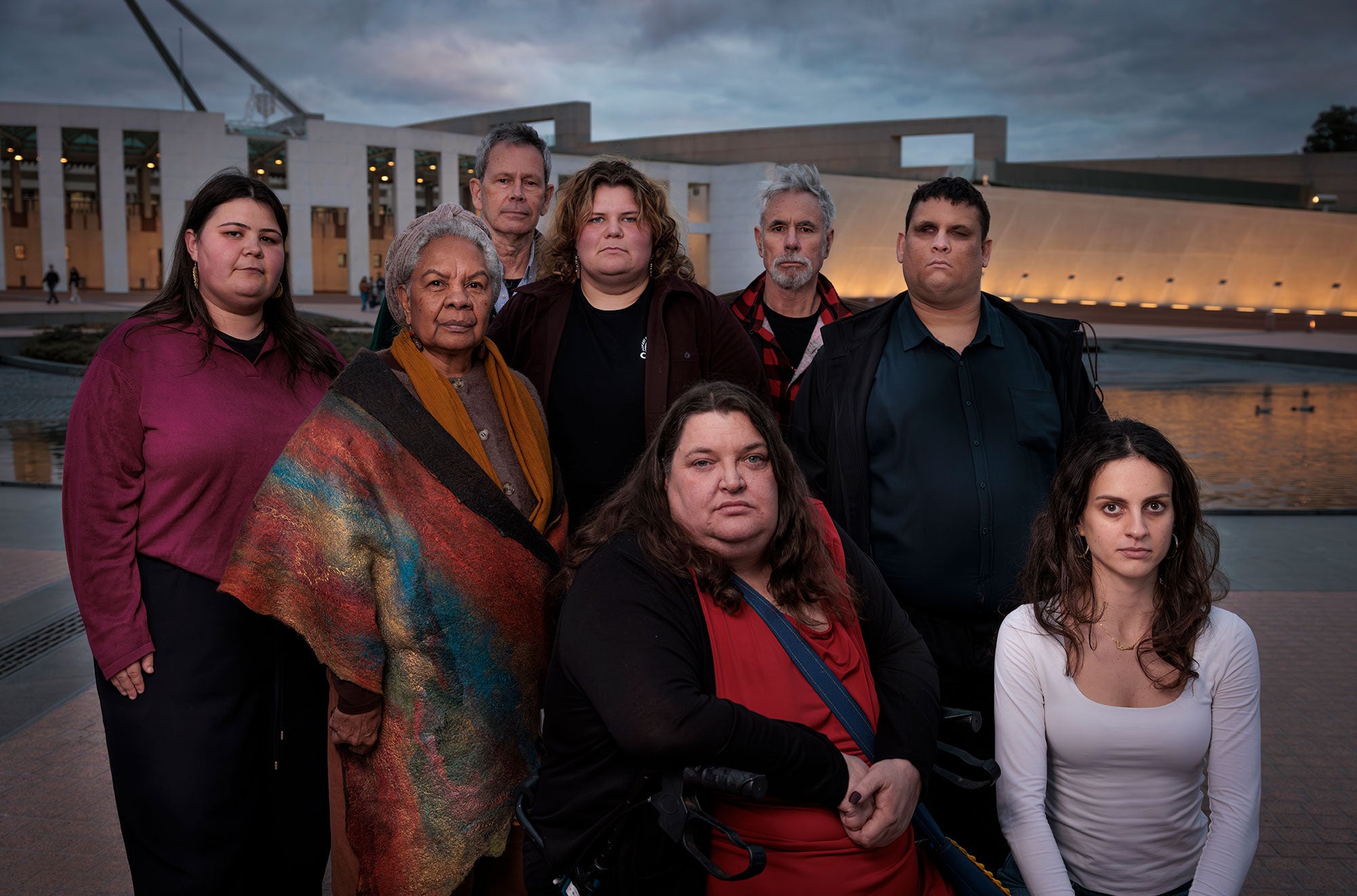 A group of 10 people, looking directly at the viewer, stand in front of Parliament House in Canberra. There are dark clouds overhead, and the soft golden lights shine up onto the front of the building from its base.