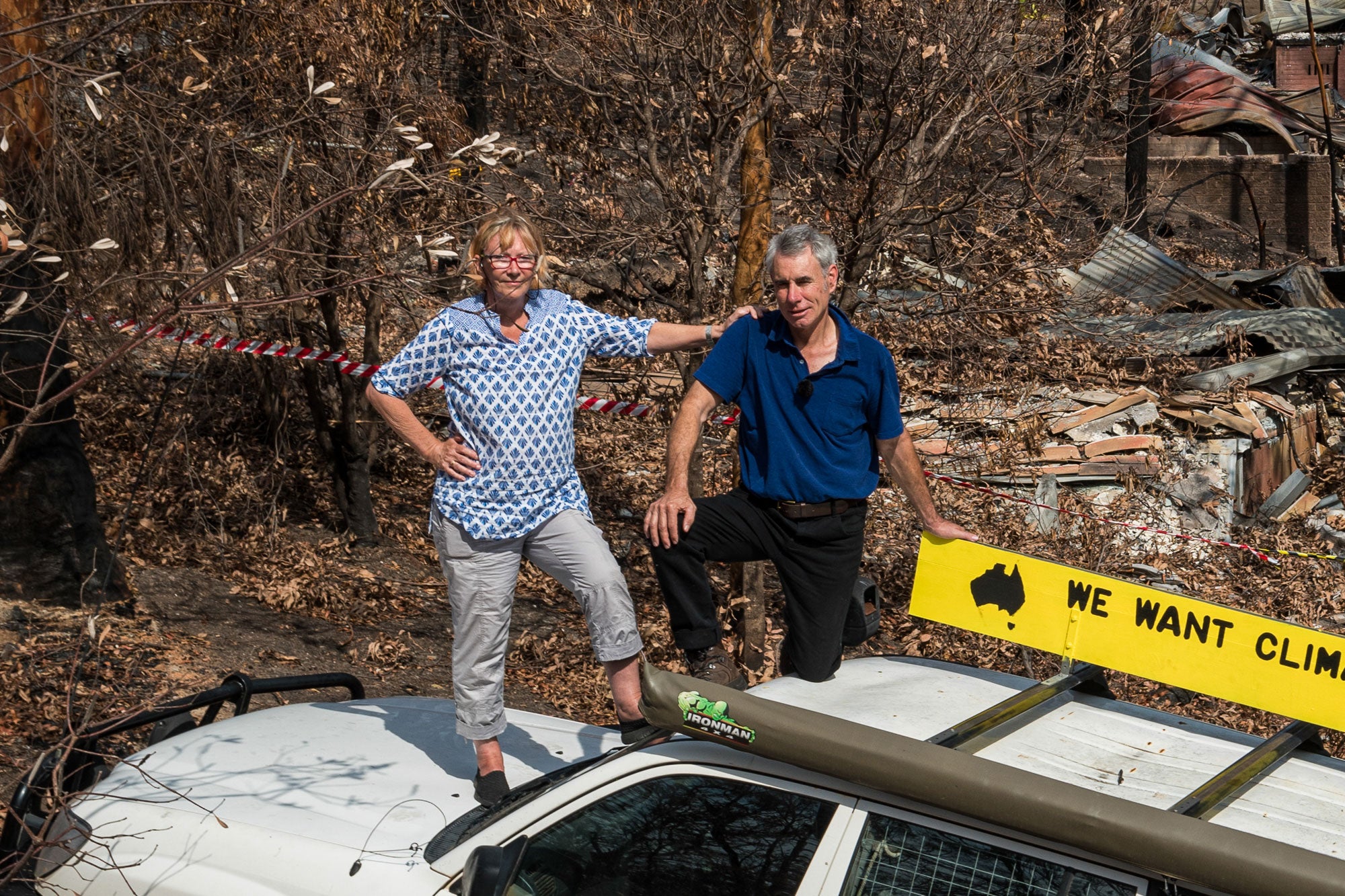 Cath and Jack, stand on top of a large white truck, with Cath standing on the front hood (left foot resting on the windshield) and Jack kneeling on the roof of the truck, with his left hand braced on a yellow sign bolted to the roof that partially reads, "We want climate ...". Cath's left hand rests on Jack's right shoulder. Behind them is a collapsed, burned out structure and a forest of burned, blackened trees. The sun is shining.