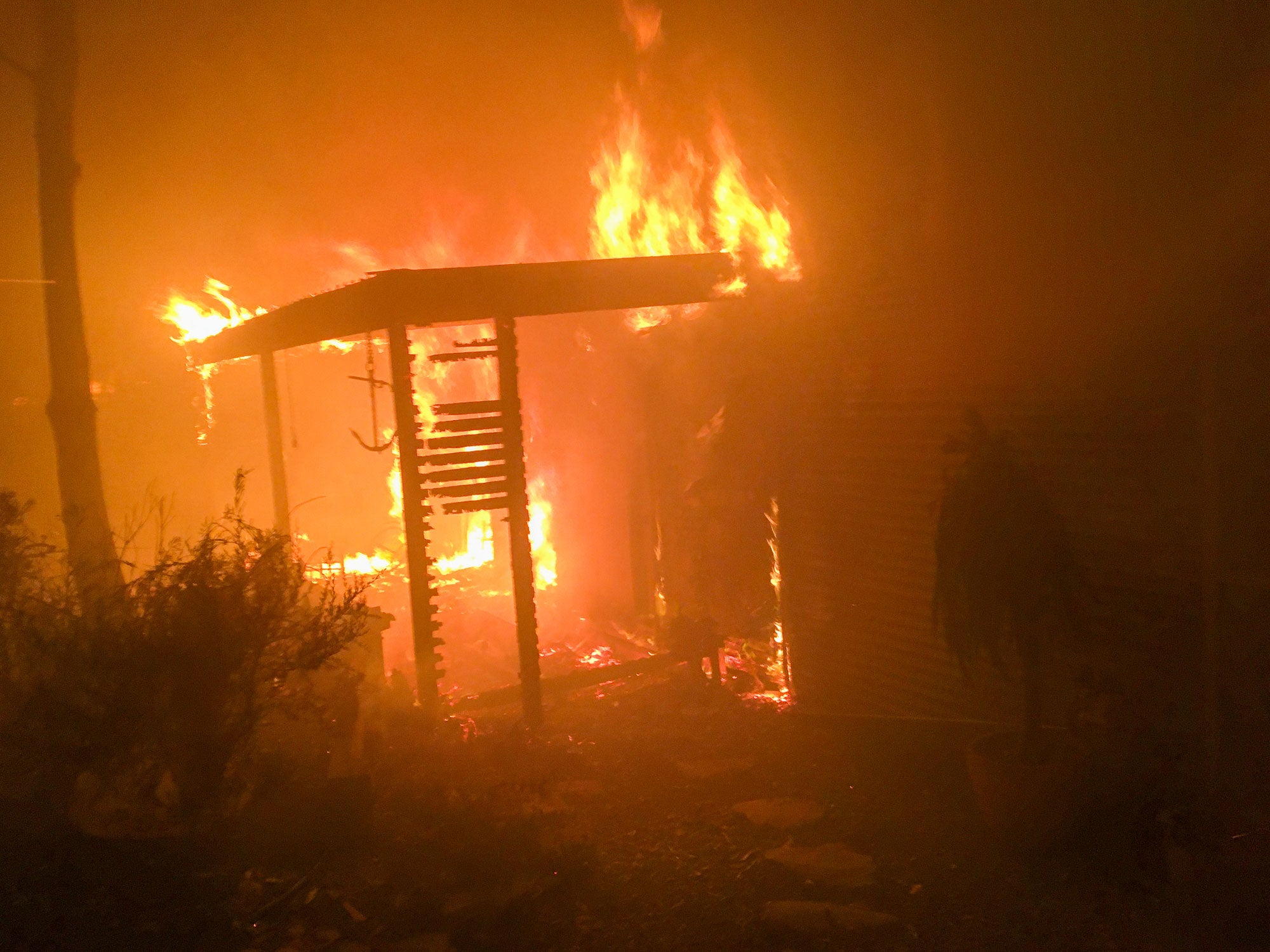 A home is engulfed in bright orange flames. An exterior veranda is on fire and partially collapsed. It is dark, and the air is thick with smoke.