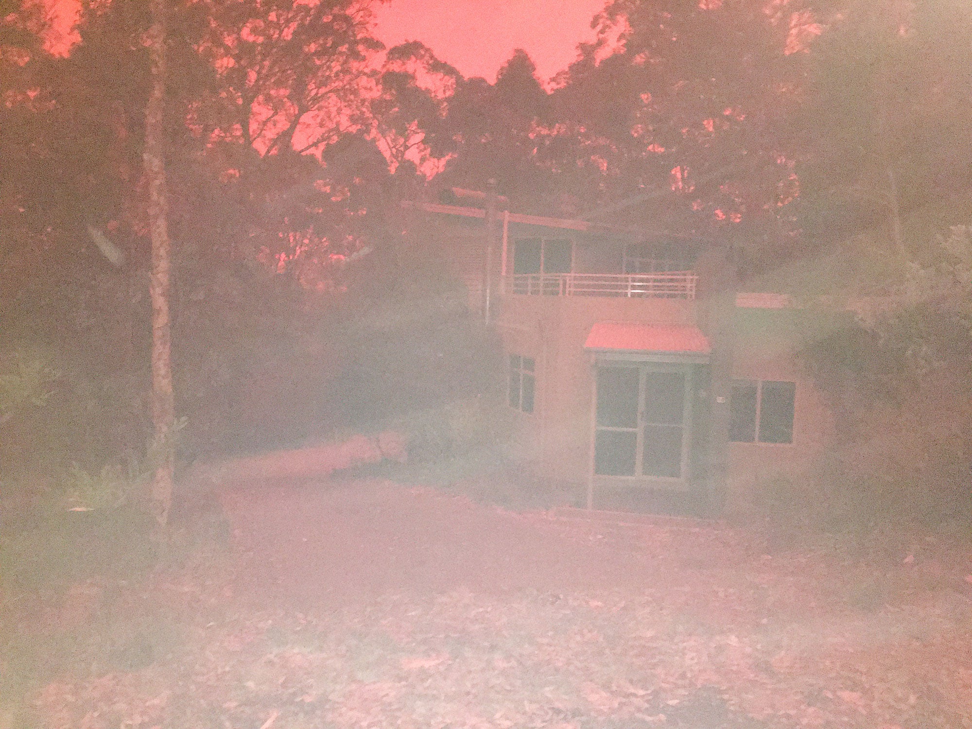 A two-story home surrounded by trees is bathed in red light. It is barely visible through thick smoke. The sky is fierce red color.