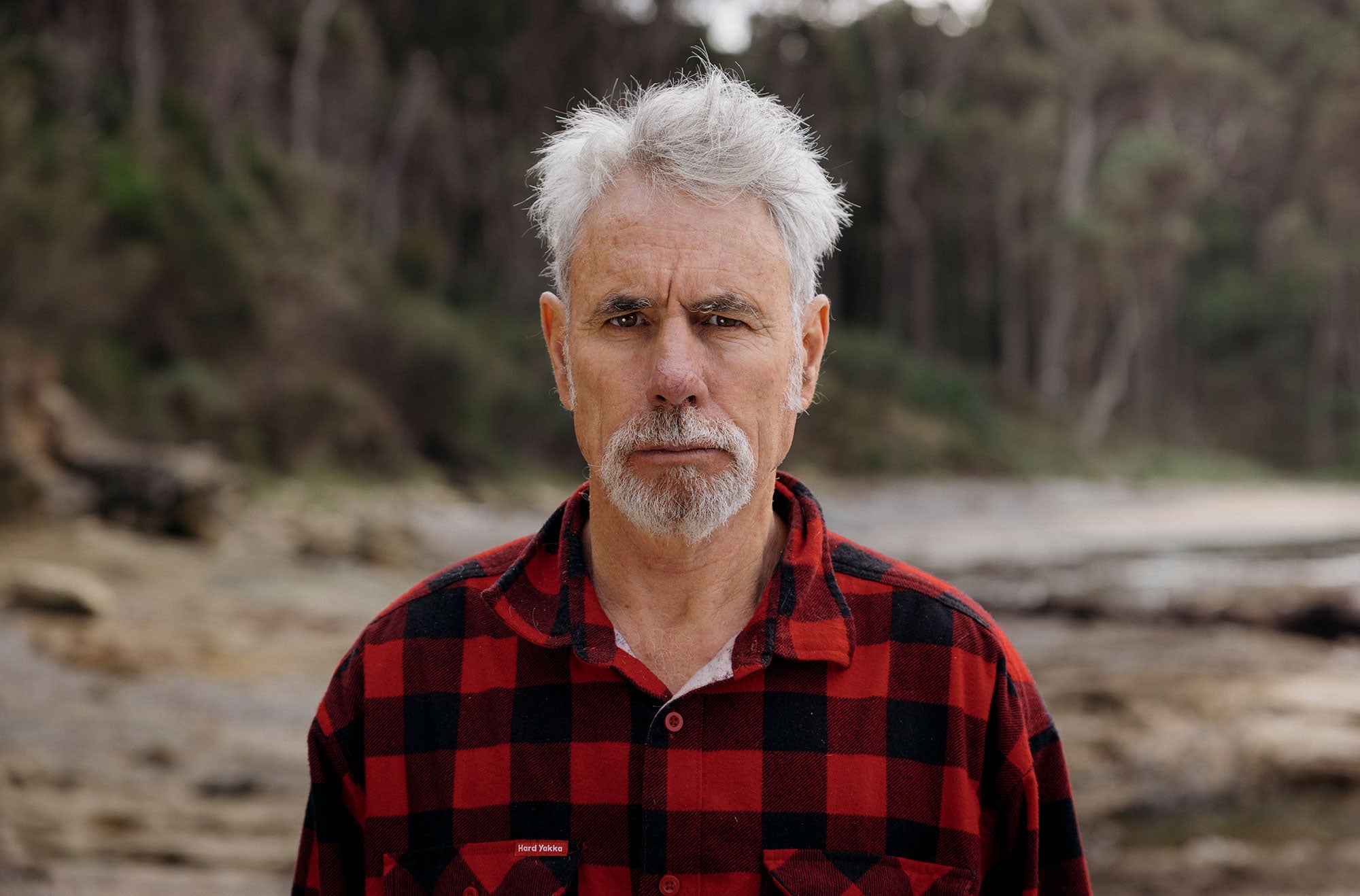 Jack stands facing the viewer unsmiling, with an intent, serious look. He is outdoors, with the background blurred brown and dark green colors. He is wearing a red and black plaid collared, buttoned-up shirt.