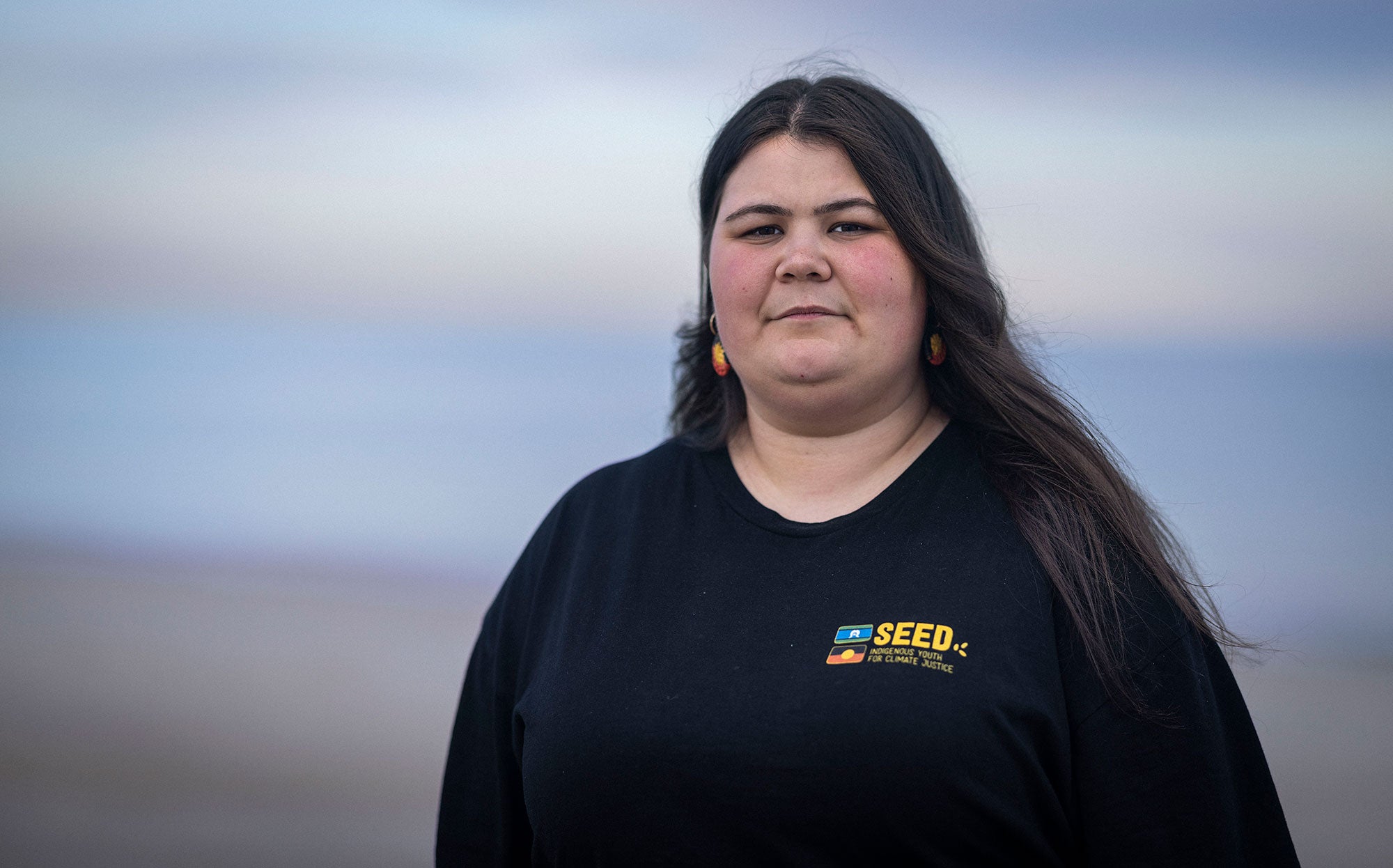 Latisha Francis stands facing the viewer with a serious expression. The photo is of Latisha from the waist up. She's wearing a black t-shirt. There is a slight hint of wind, as her long, black hair appears to be waving in the wind. The background is a blurred bluish-purple color.