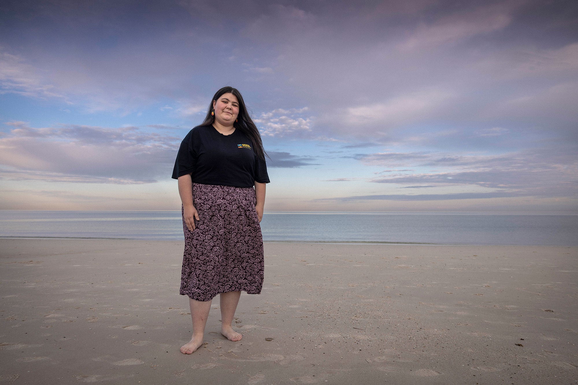 Latisha is standing barefoot on a sandy beach, with the ocean behind her at a safe distance. She looks slightly down towards the viewer with a serious smile. Her arms are at her side. She is wearing a black t-shirt and a long, flowing dark purple floral print skirt. The beach is bare and serene. The sky is a blue-ish pink with purple clouds.