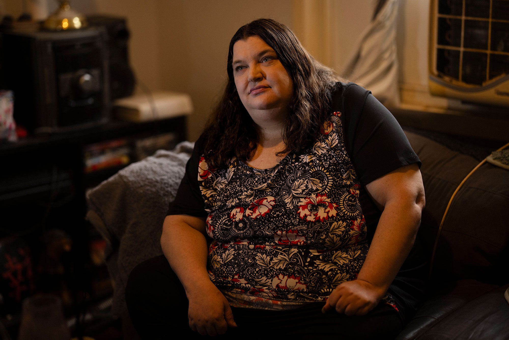 Mel sits on a sofa inside her home, with her hands lightly braced against her thighs. She looks slightly to the side of the viewer, with a thoughtful expression.