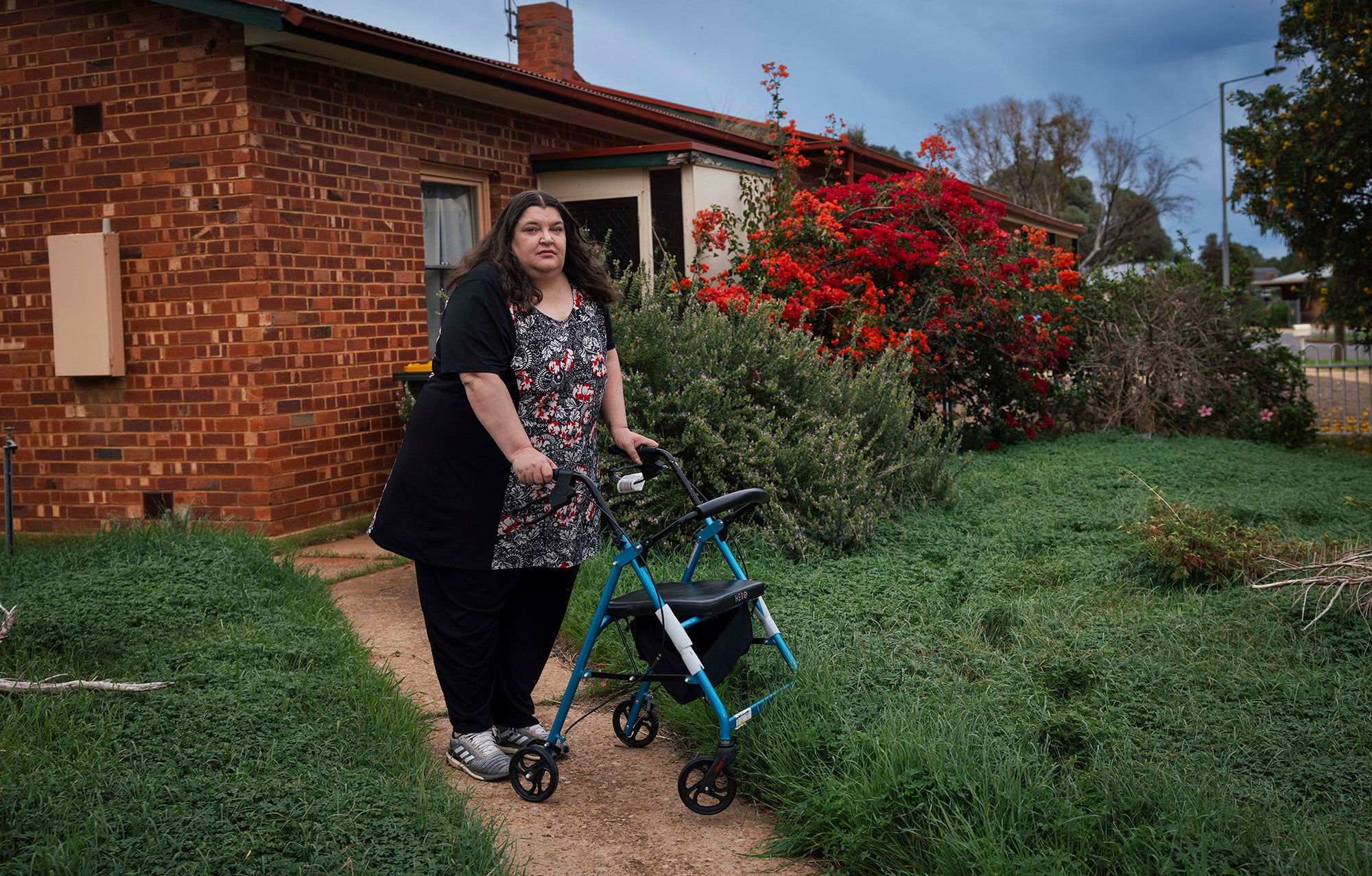 Mel stands on a dirt garden path in front of her brick home, leaning against her rolling walker, looking at the viewer with a serious expression. Bright green groundcover grows lushly on either side of the garden path. Flowering bushes frame the front of the house. The sky overhead is blue, and a few other homes are visible to the far right.