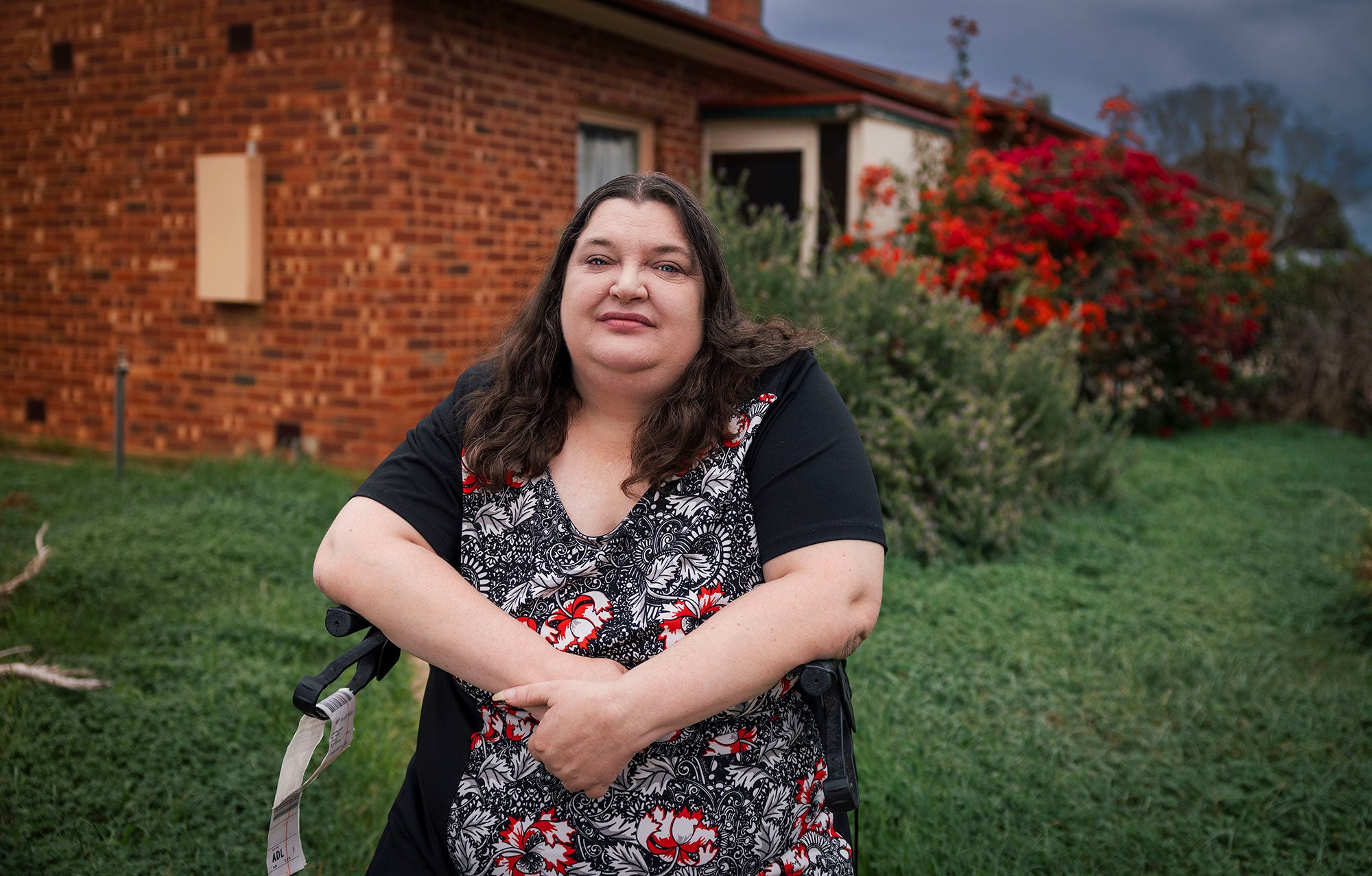 Mel sits in a wheelchair outside of her home with her hands clasped in front of her. She looks directly at the viewer with a serious expression in her eyes. The brick house is surrounded by a lush garden, with rich green growth, and bushes with bright red flowers.