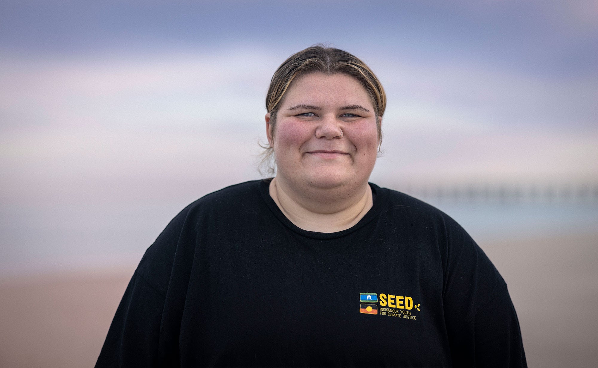 Pam stands with a friendly smile, facing the viewer. The background is out of focus, with a long pier just visible. She's wearing a black t-shirt.