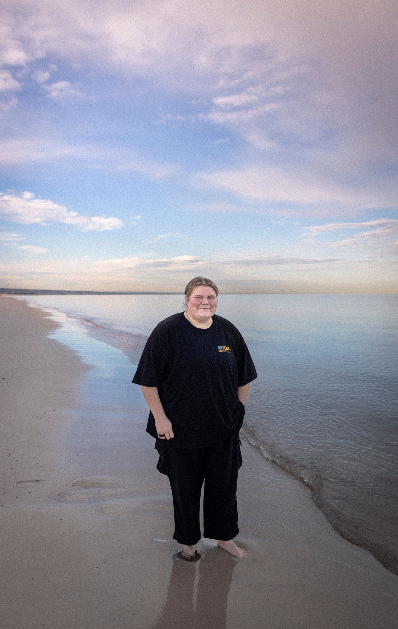 Pam Francis stands smiling on a beach, looking at the viewer, with her feet in the wet sand, and the ocean tide gently flowing in or out. She is wearing black pants and a black t-shirt. One of her hands is in her pocket, and the other is by her side. There are a few gentle white clouds in the bluish-pink sky.