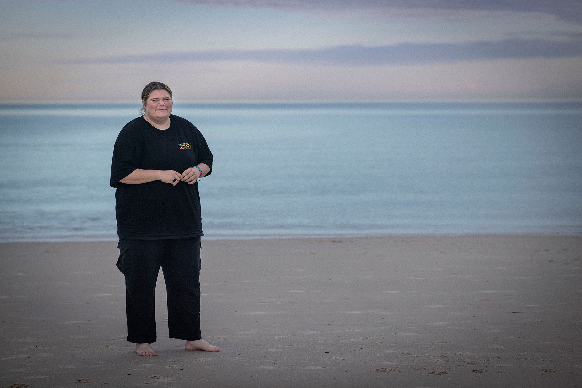 Pam Francis stands barefoot on a beach, smiling at the viewer. The ocean is behind her at a safe distance. The ocean looks calm. She is wearing a black t-shirt and black pants, and colorful bracelets on her left wrist. The sky is a bluish pink.
