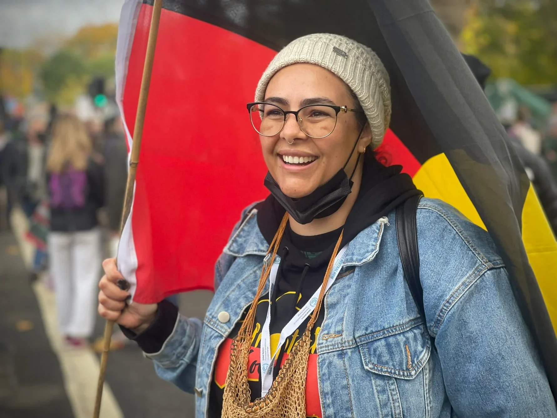 Rikki stands with an open-mouth, friendly smiled, holding a flag with red, white, yellow, and black colors. The flag is flowing behind her. She is looking slightly off screen. There are many people (out of focus) around her. She's wearing a black face mask that is pulled down below her chin. Rikki is wearing stylish large glasses, a knit beige hat, a jean jacket, and a black t-shirt with design.