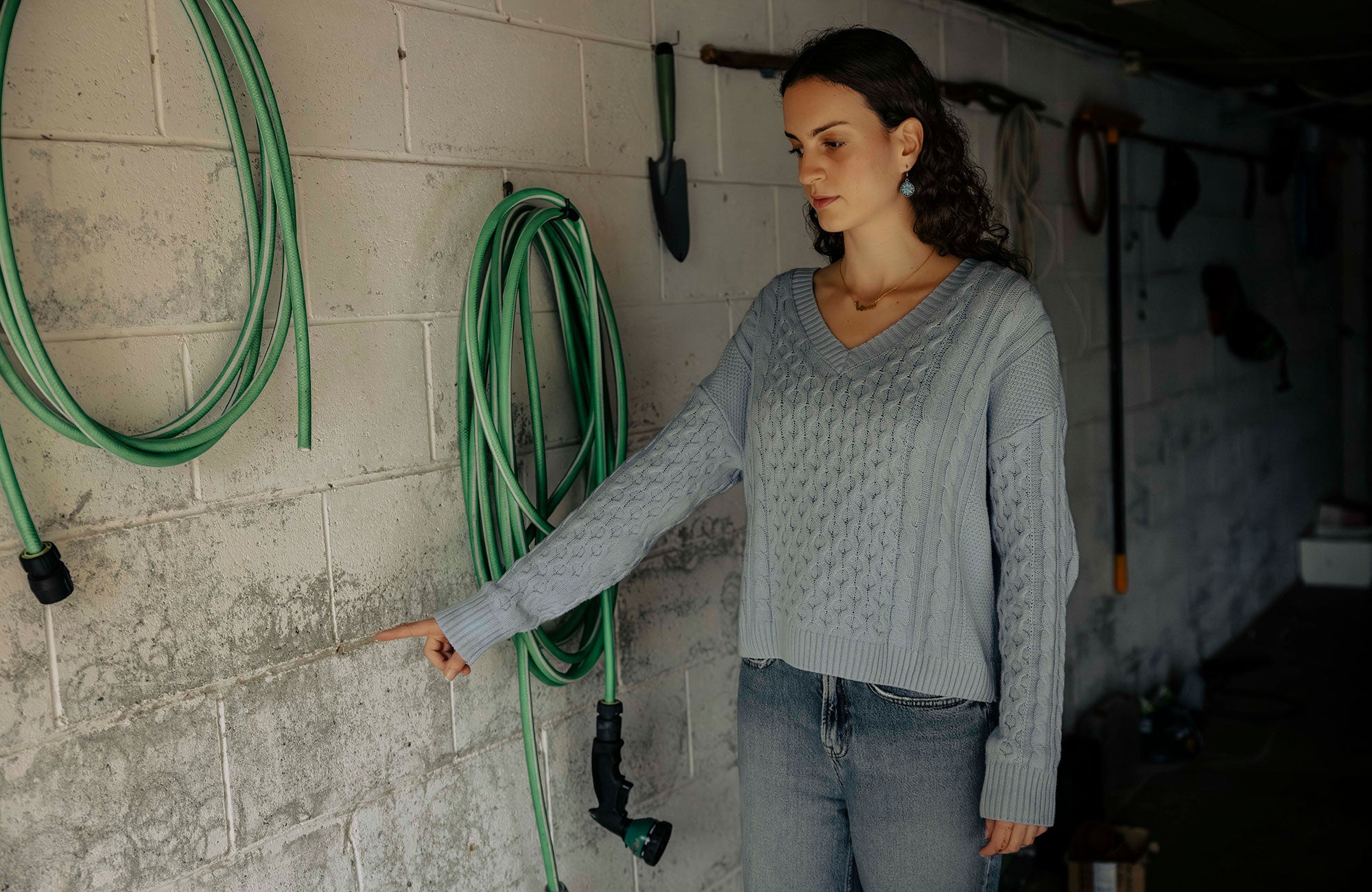 Sama, wearing a light blue sweater and jeans, stands inside her family home's garage, pointing to the place on the grey cinderblock wall where floodwaters reached, at a height that appears to be about three feet. Two neatly coiled green garden houses hang on the wall where she is pointing. Other gardening tools are hanging on the wall behind her.
