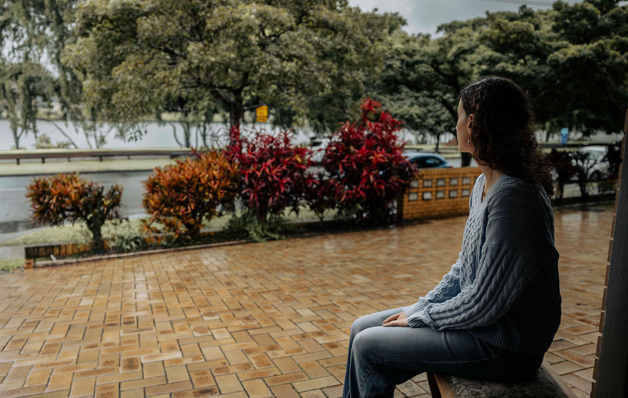 Sama sits on a bench outside of the garage of her family home. She is partially turned away from the viewer, looking at the Brisbane River which lies a few feet away, just past a brick-paved path and a road. Trees line the river bank. It is a wet and rainy day.