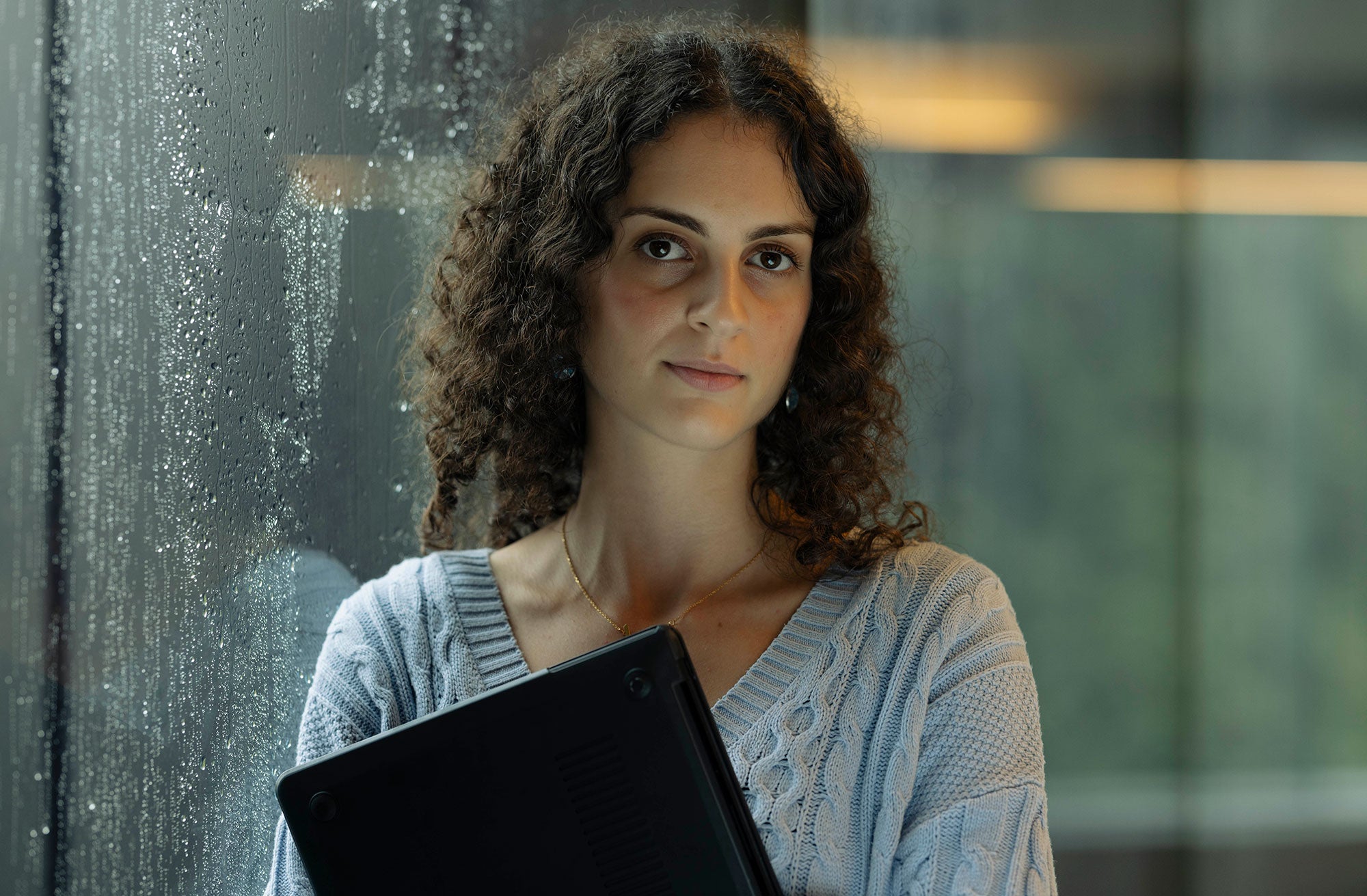 Sama stands leaning against a floor-to-ceiling glass window, holding her closed black laptop in front of her chest and looking straight at the viewer with a serious, strong expression. Water droplets from a recent or ongoing rainfall are visible on the outside of the glass window.