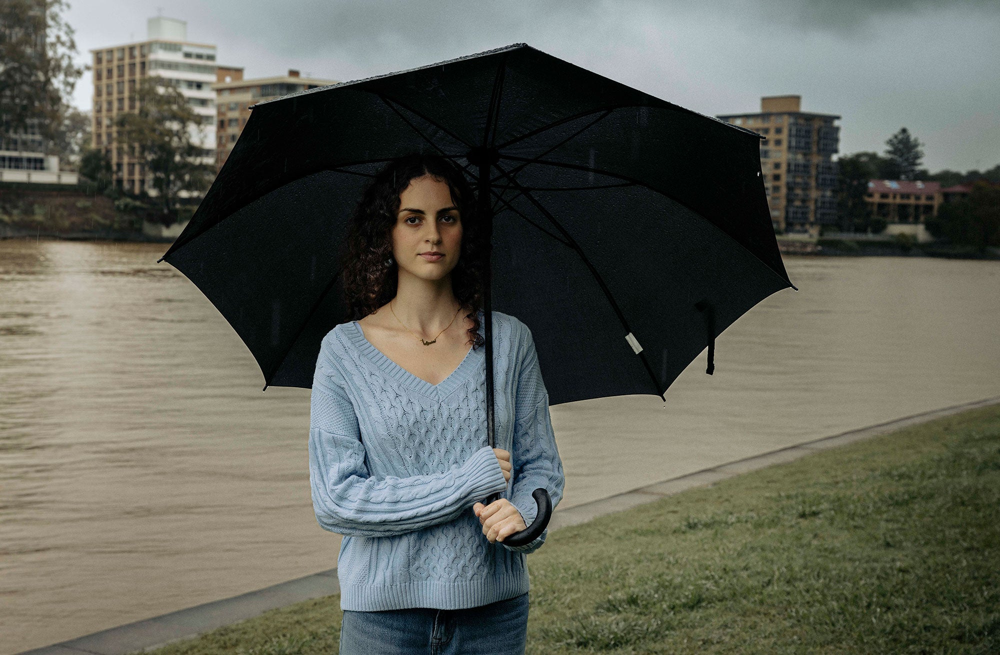 Sama stands in a grassy area on the banks of the Brisbane River, holding a large black, opened umbrella to shelter herself from the rain. She is directly facing the viewer with a serious expression. The Brisbane River is brownish color and is almost reaching the grassy area. The day is overcast. Buildings are visible on the opposite bank of the river, behind Sama.