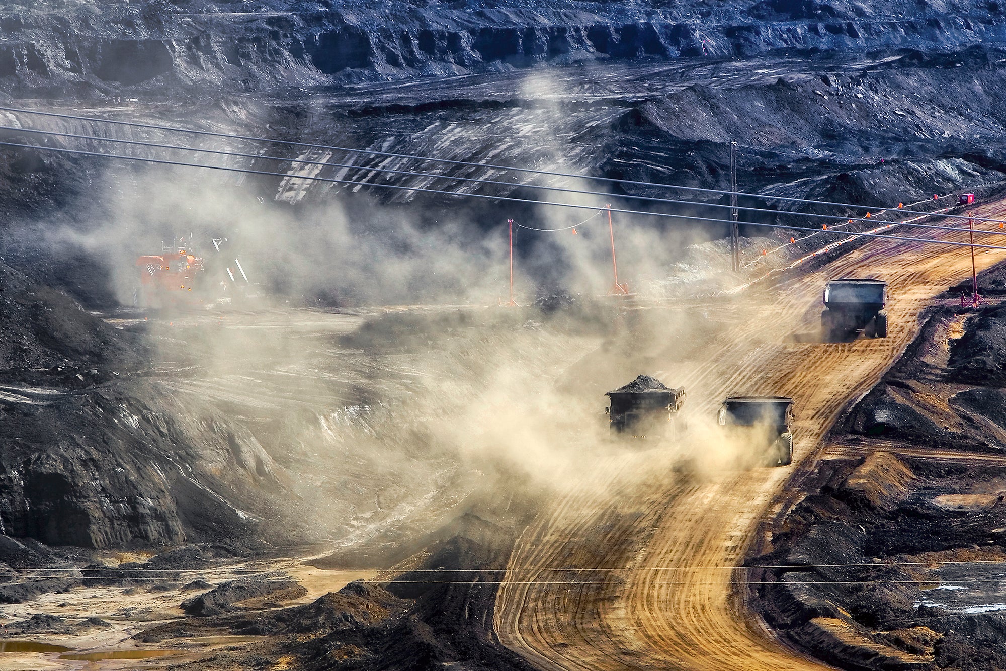 A photo showing black, terraced earth with a dirt road going through it. There are three dump trucks driving on the road kicking up a lot of dust.