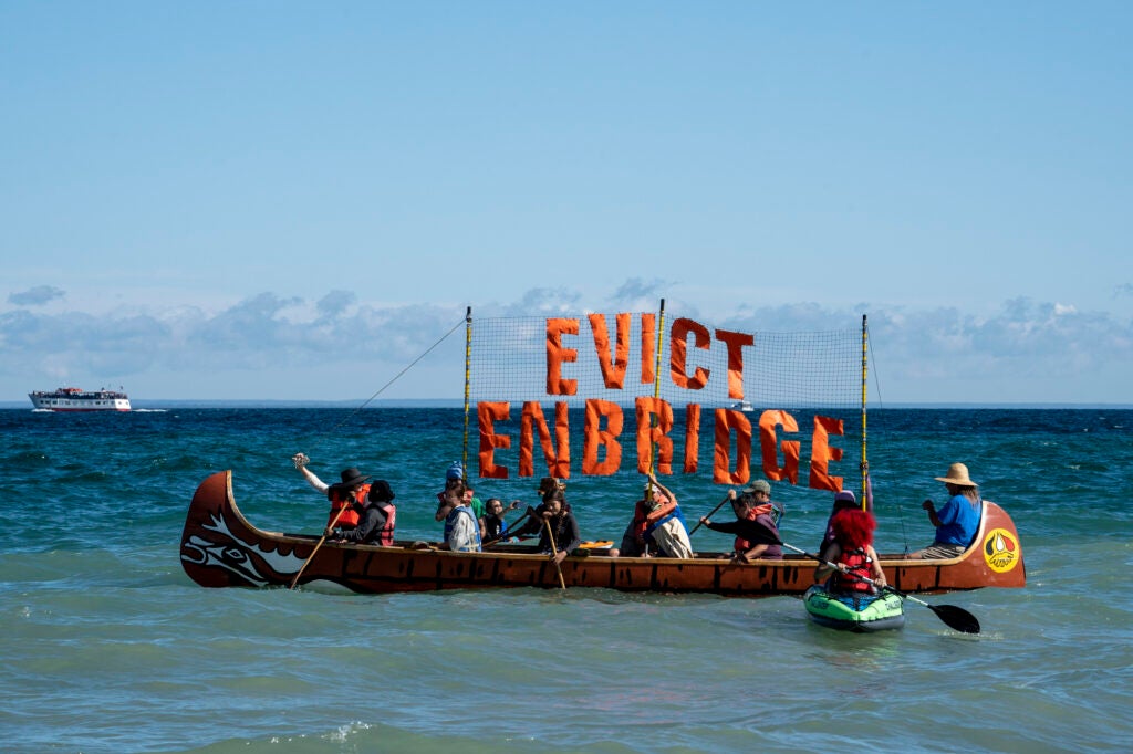 A canoe with about a dozen people in it on a large lage, holding a large sign that says "Evict Enbridge"