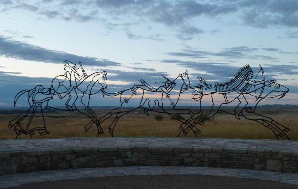 A bronze sculpture silhouetted against the sky of three Native American warriors riding to battle, and a woman handing off a shield to one as she watches them ride off seemingly into the sky, where the spirit world dwells according to traditional tribal beliefs. A faint pink sunset and darkening clouds are visible through the outlines of the sculpture. A low curved, dual level stone wall is visible below the sculpture.