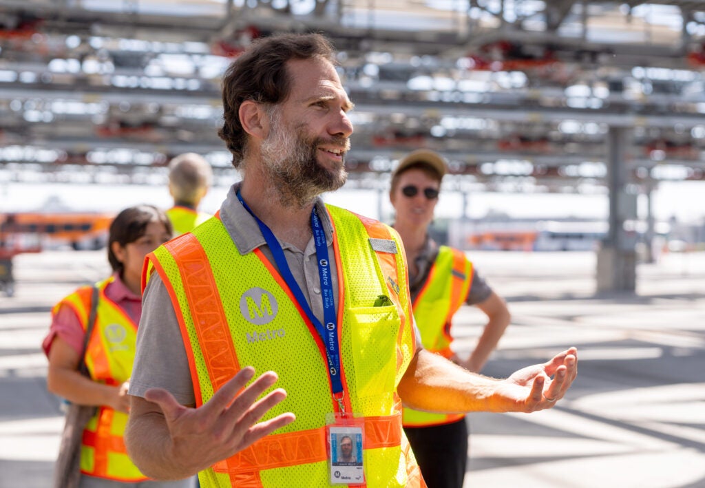A man in a yellow reflective vest talks with people under a metal structure.