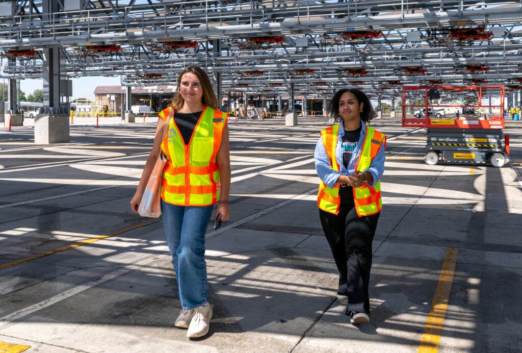 Two women walk under a metal structure at a bus depot.