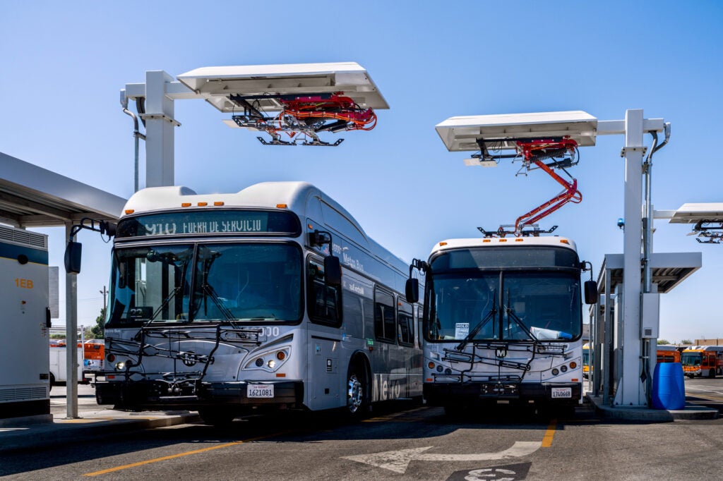 Two buses parked under large charging devices above them, which are raised up by metal poles.