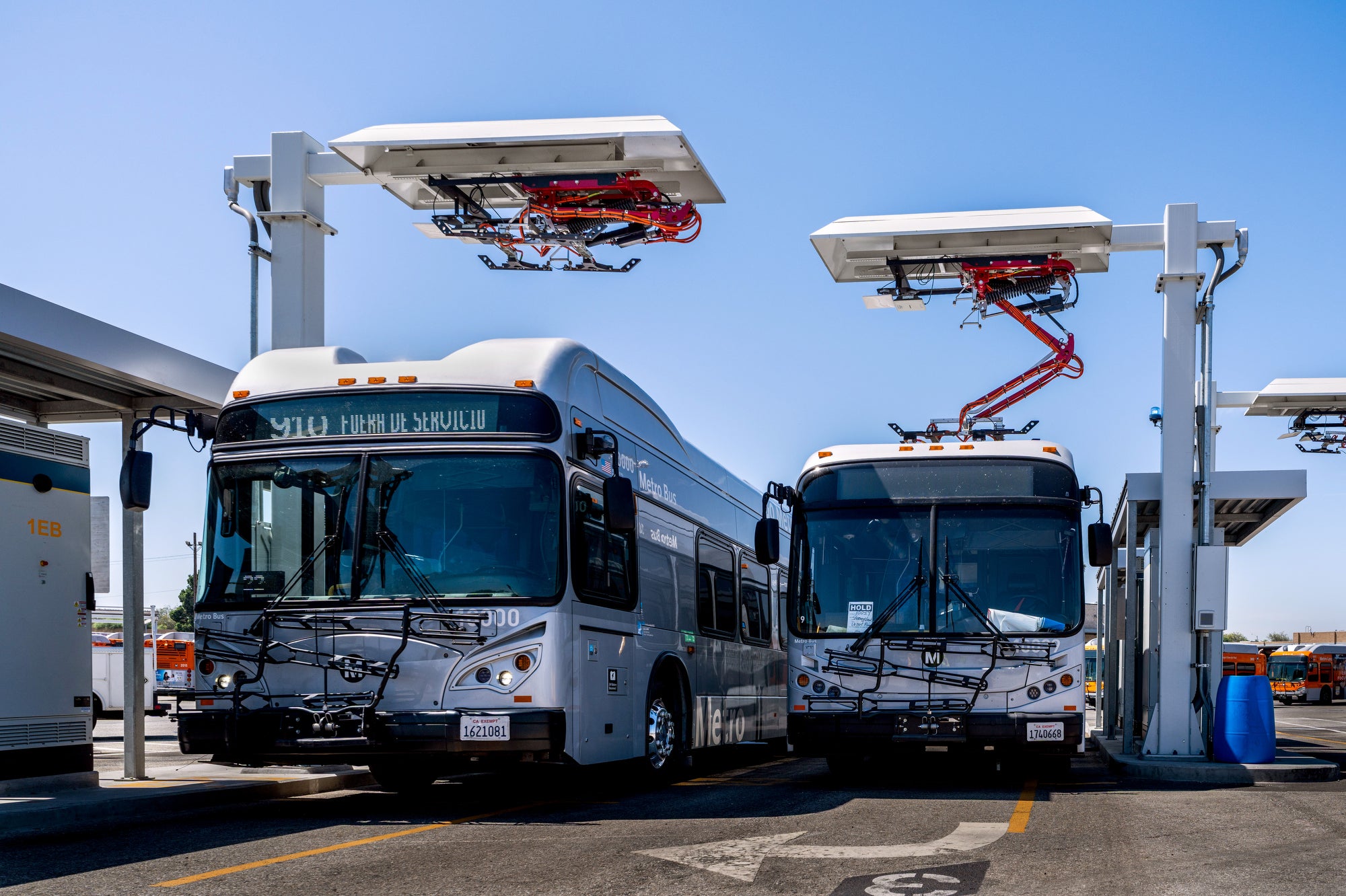 Two buses parked under large charging devices above them, which are raised up by metal poles.