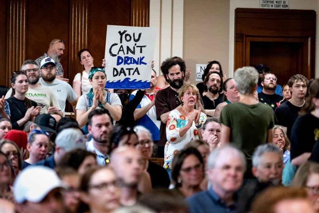 A large crowd of animated, serious-looking people pack a courtroom, with some clapping and standing. One person holds up a large sign that reads, "You can't drink data" written above a drawing of blue waves.