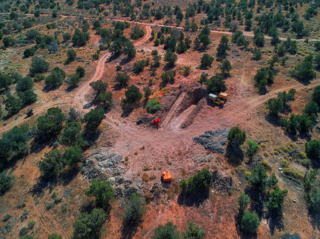 An aerial view of a mining shaft. Bright green trees dot a red-brown landscape. A few construction equipment and containers are visible near the mining shaft.