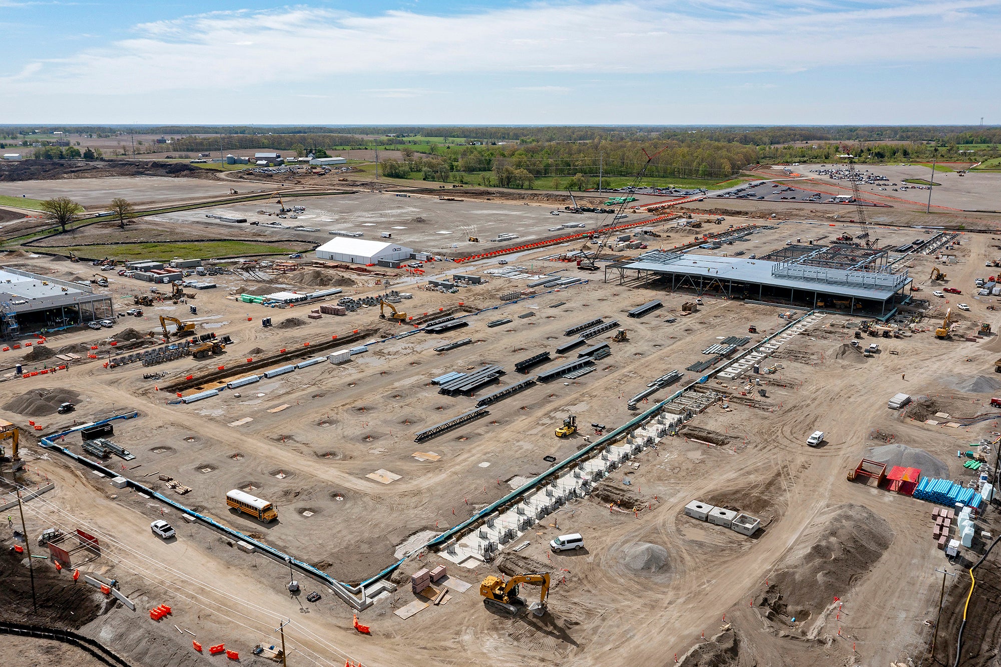 A large, graded, dirt construction site with machinery, vehicles and some buildings under construction, with flat farmland and trees visible in the distance.