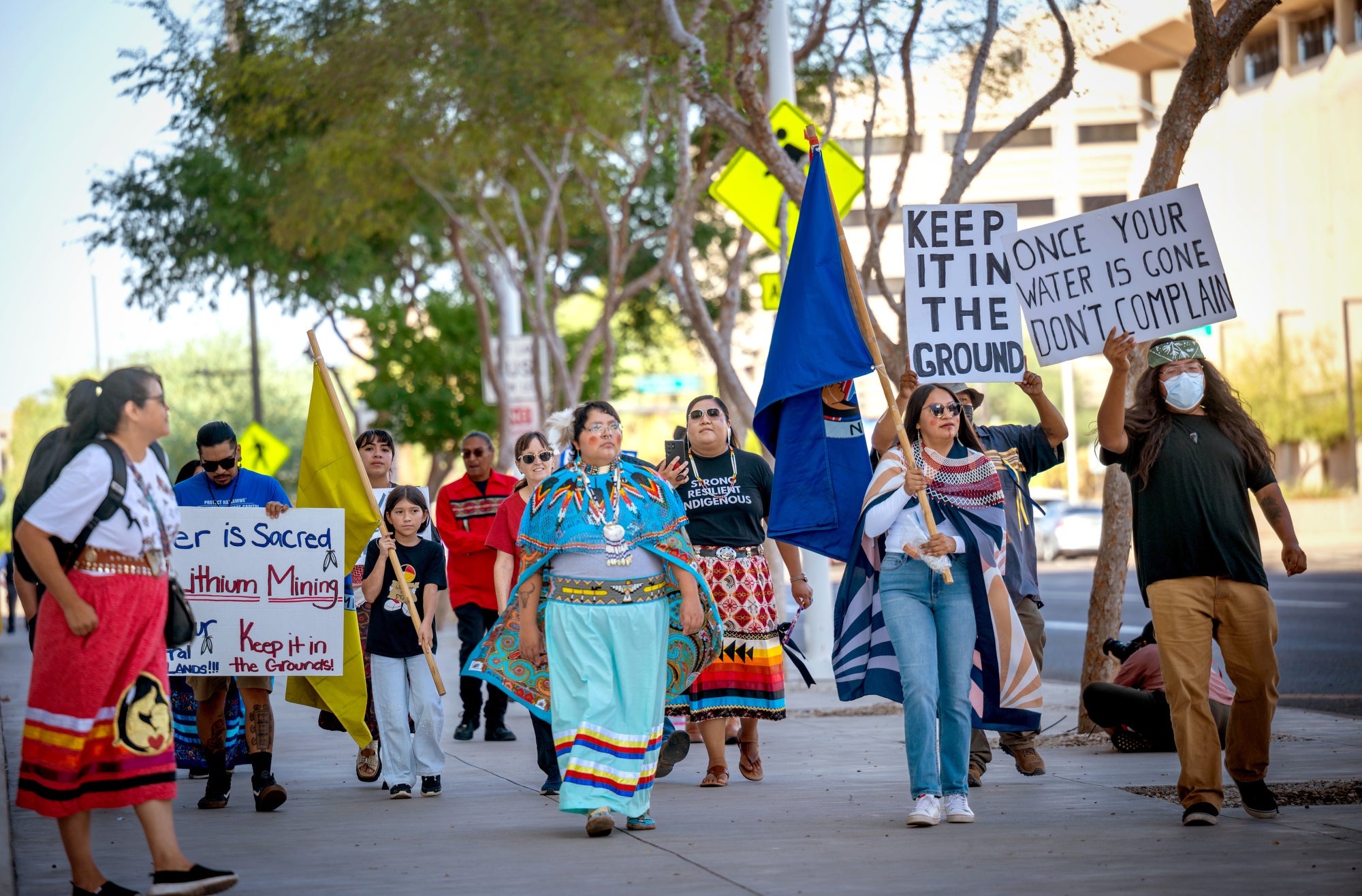 Members of the Hualapai Tribe march on a city sidewalk holding flags and signs. Some of the signs say "Keep it in the Ground" and "Once Your Water is Gone Don't Complain"