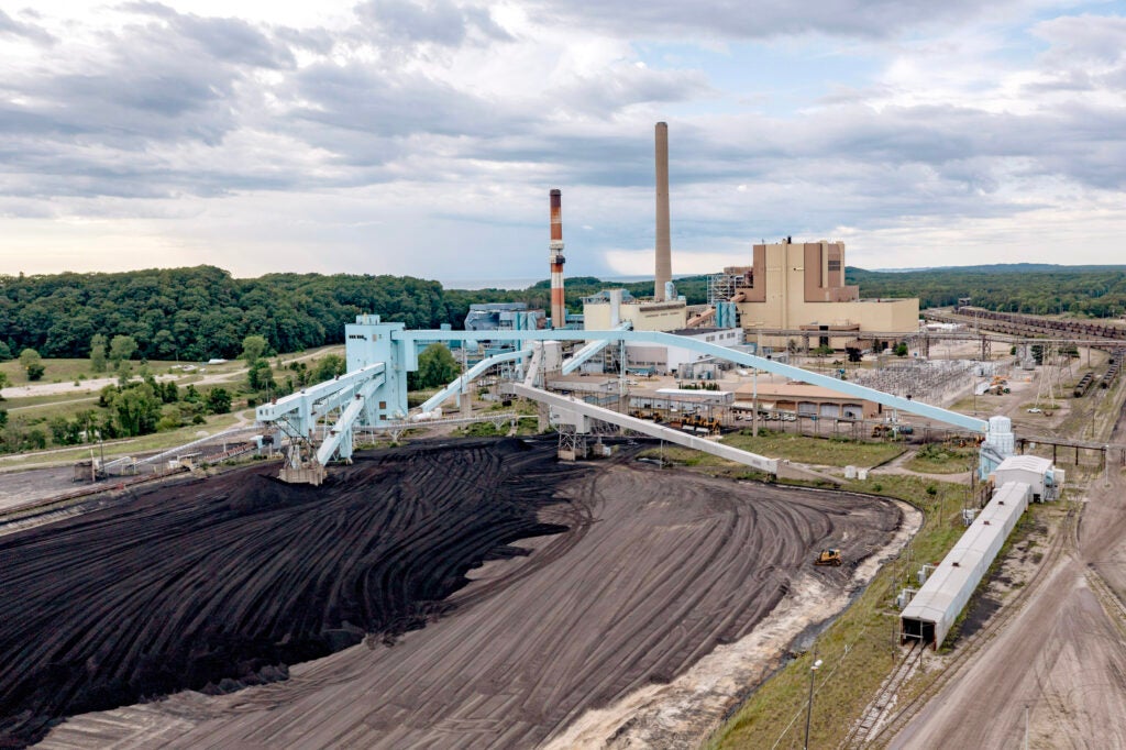 Aerial view of the J.H. Campbell coal-fired power plant, showing large coal storage piles, conveyor systems, smokestacks, rail lines, and industrial buildings surrounded by forest under a cloudy sky.