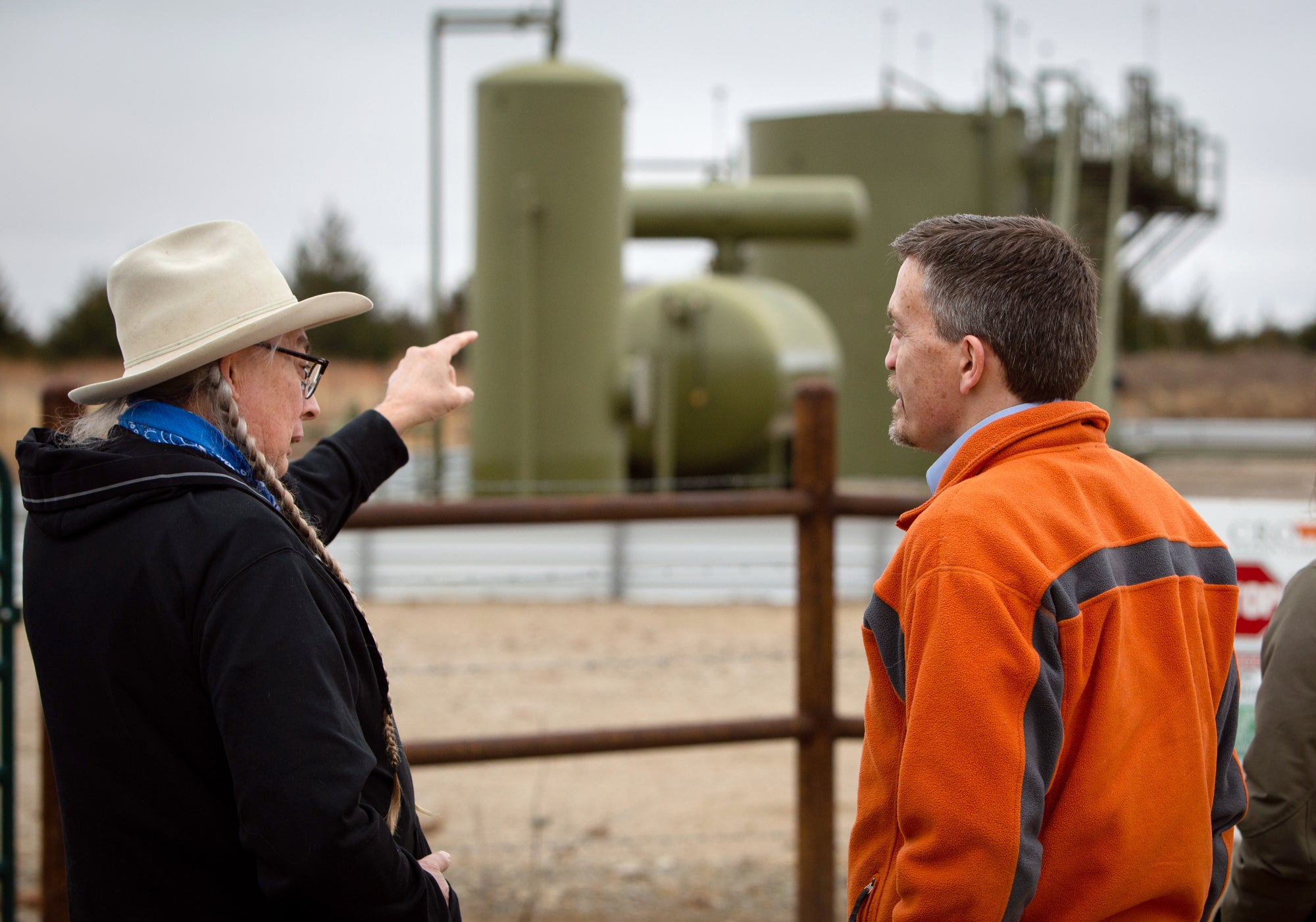 Two men stand near a metal fence and the one on the left is talking and pointing to machinery, out of focus in the background.