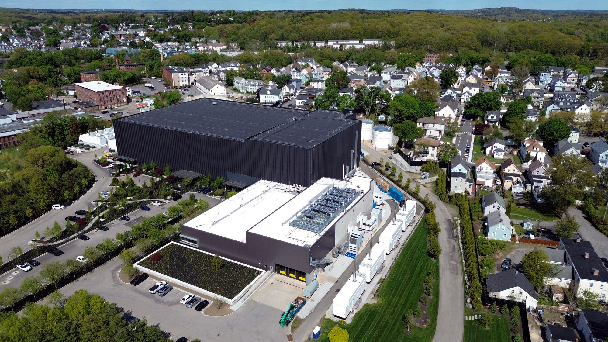 An enormous black, boxy-shaped building towers over and dwarfs a cluster of residential homes. A single road appears to separate the large industrial complex from the homes. Beyond the homes is a dense, green forest.
