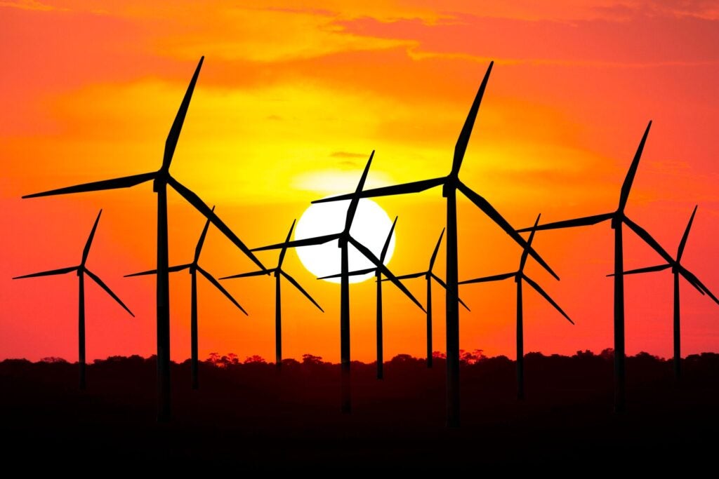 Silhouettes of multiple wind turbines against a bright orange and yellow sunset, with the sun low on the horizon behind the turbines.