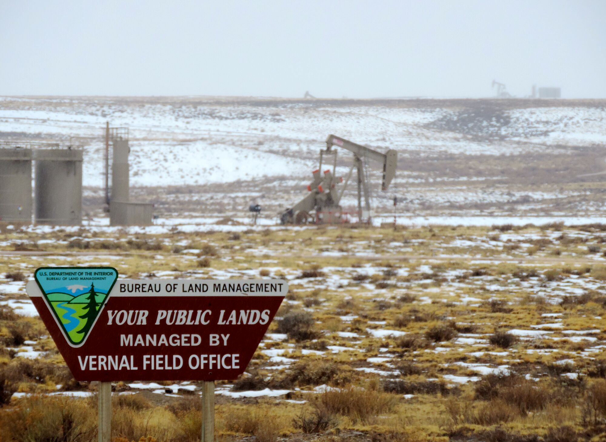 A Bureau of Land Management sign is in the foreground, with an oil derrick behind it.