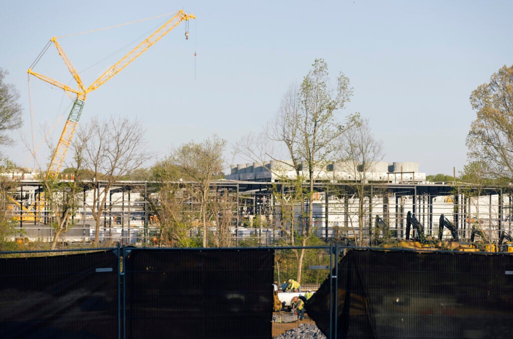 A long, wide construction site dominates the middle of the frame, with a crane above it. In the foreground on the lower fourth of the picture, there is a fence covered with a black tarp. There is a gap in the middle of the tarp showing a crew of construction workers at work.