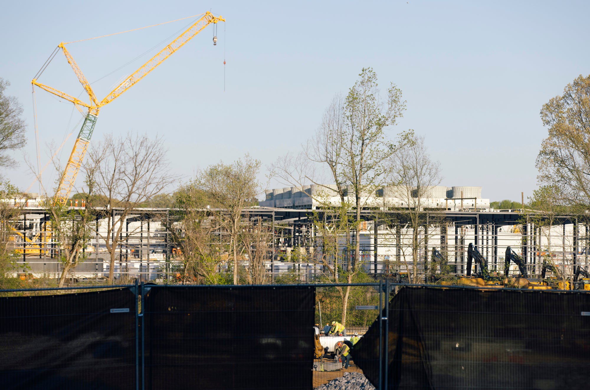 A long, wide construction site dominates the middle of the frame, with a crane above it. In the foreground on the lower fourth of the picture, there is a fence covered with a black tarp. There is a gap in the middle of the tarp showing a crew of construction workers at work.