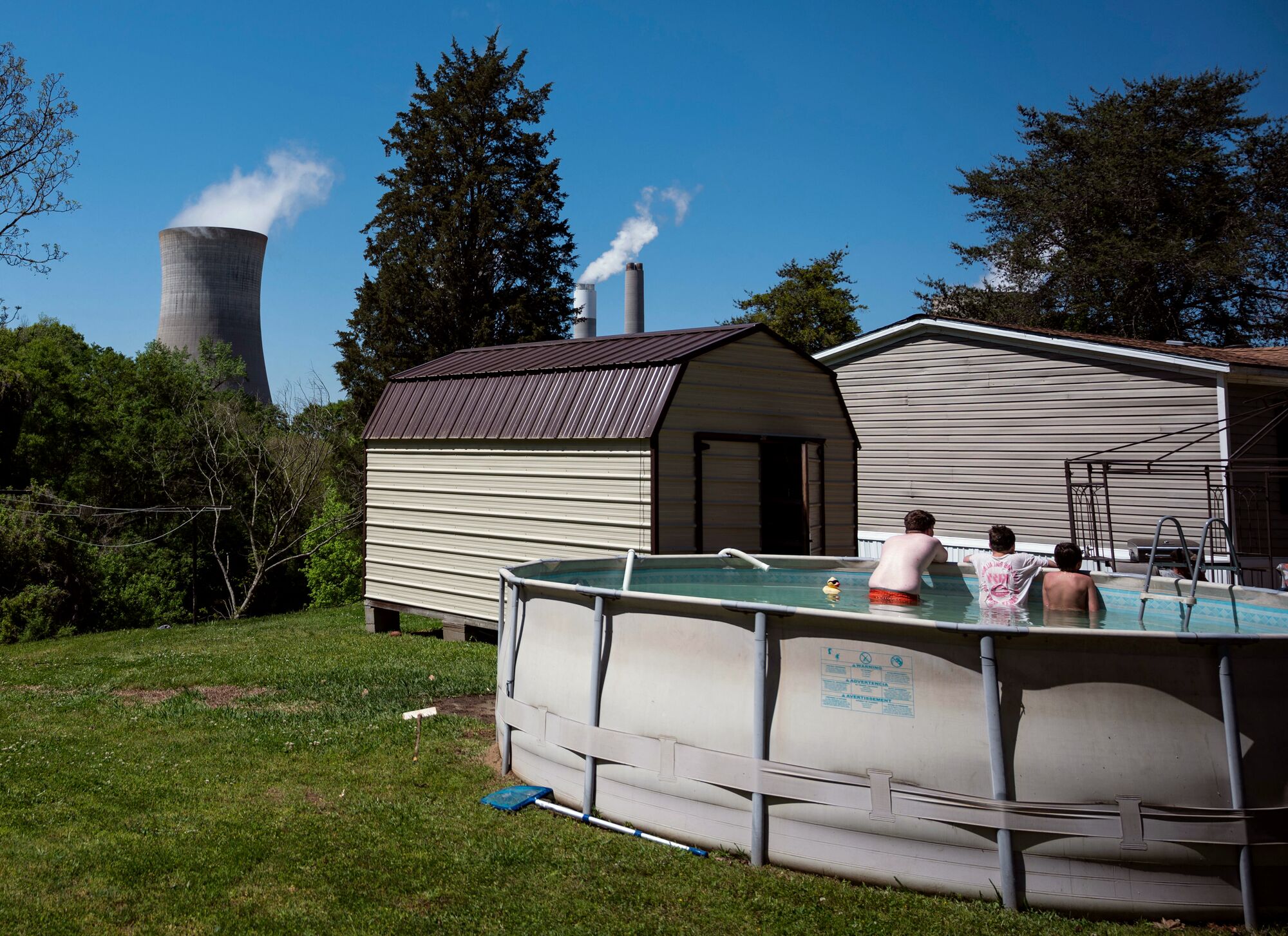 Kids lounge in an above-ground swimming pool in a backyard, with their backs to the viewer. They are looking at two enormous smokestacks that tower over the residential buildings. Wispy white smoke is coming out of the smokestacks. The sky is a cloudless, brilliant blue.