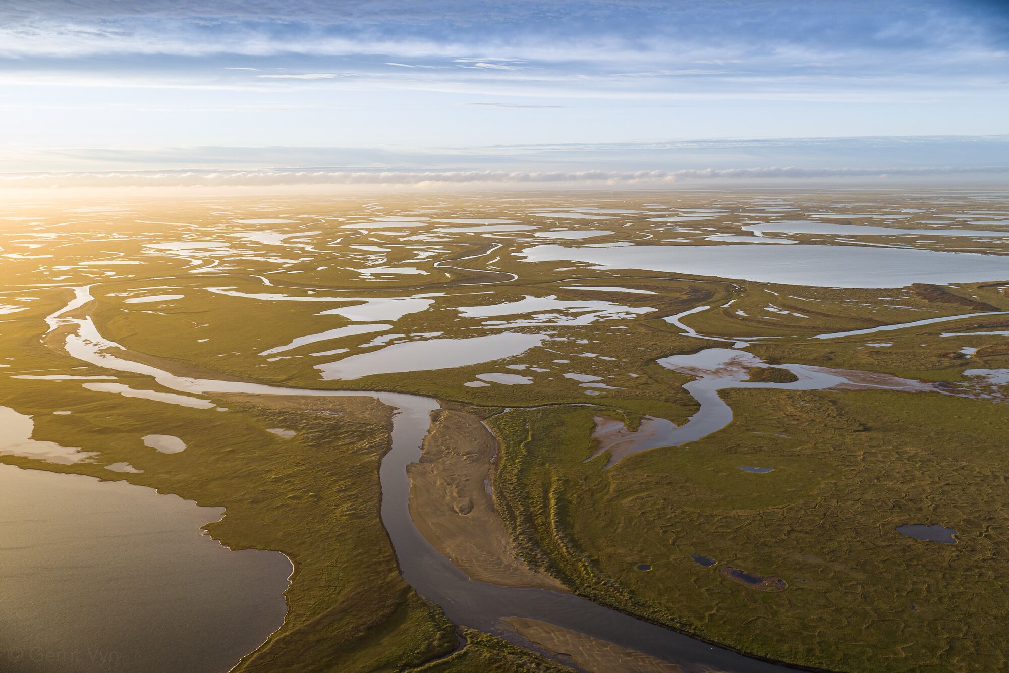 Aerial view of wetlands, lit by the glow of the sun from off to the left. Numerous pockets of water dot the landscape as far as the eye can see.