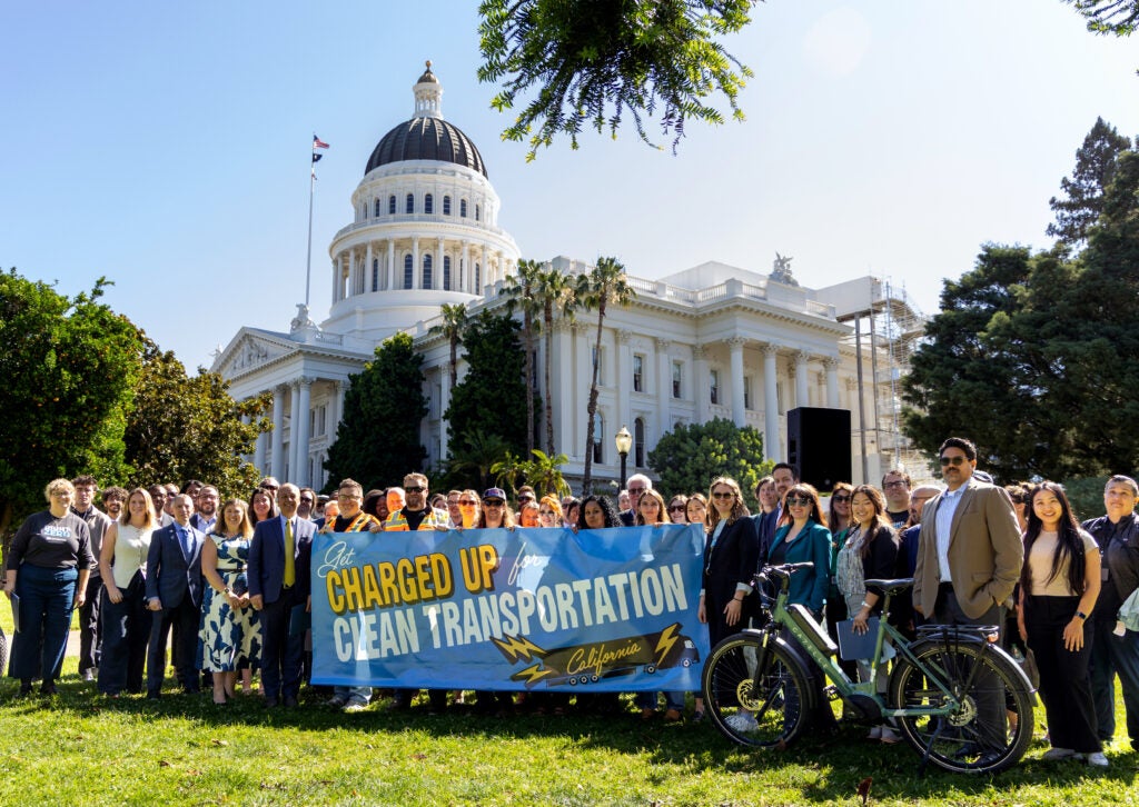 A large group of smiling, cheerful people are lined up together on a grassy field, holding a huge banner that reads, "Get Charged Up for Clean Transportation, California!" There is a an electric bike visible in front of the front row of people. The majestic dome and the grand white columns of the Capitol building tower over them. It is a cloudless, sunny day.